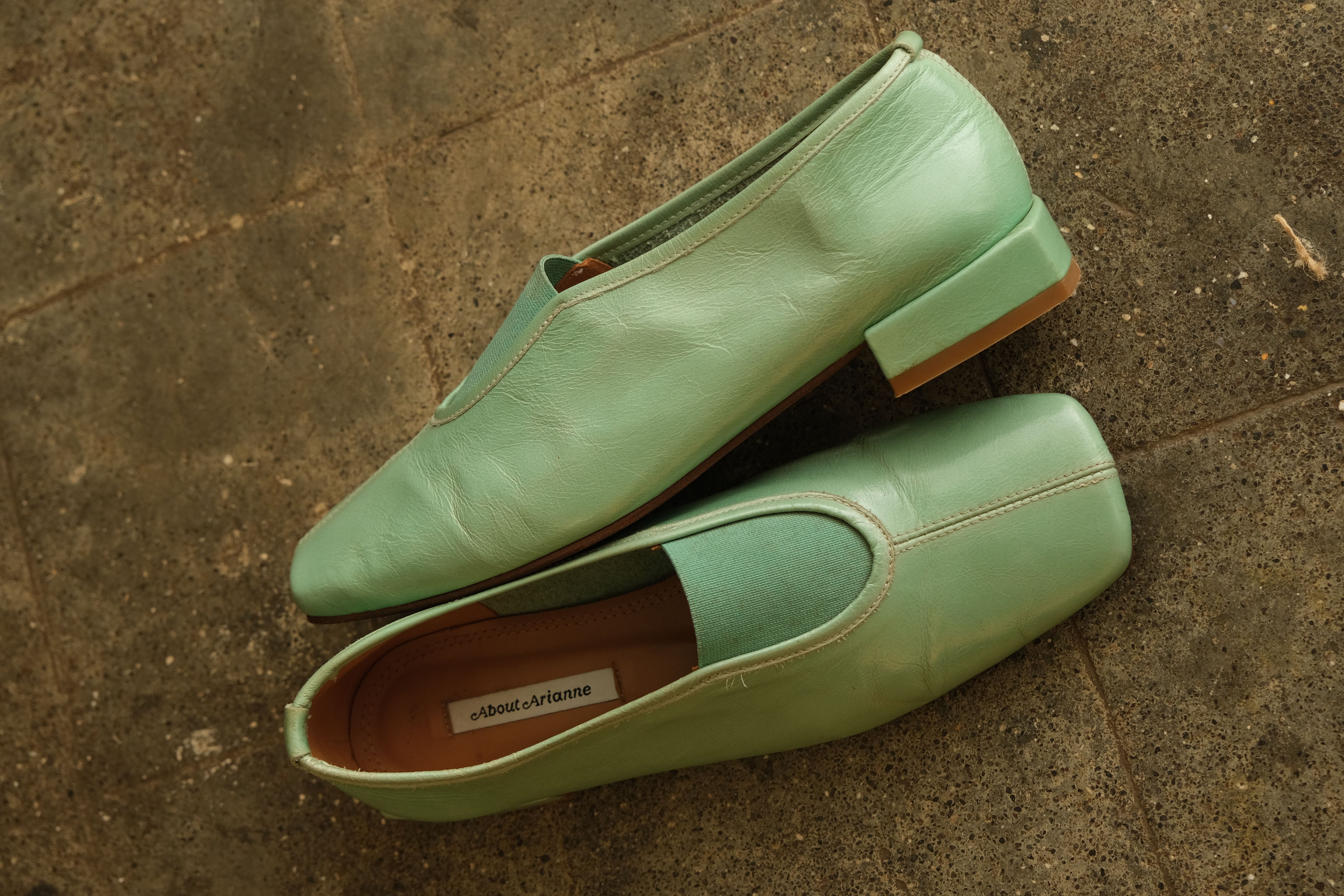A pair of mint green leather slip-on shoes with a low block heel, displayed on a textured concrete surface. One shoe is positioned upright, showing its side profile and heel, while the other lies on its side, showing the open toe and interior.
