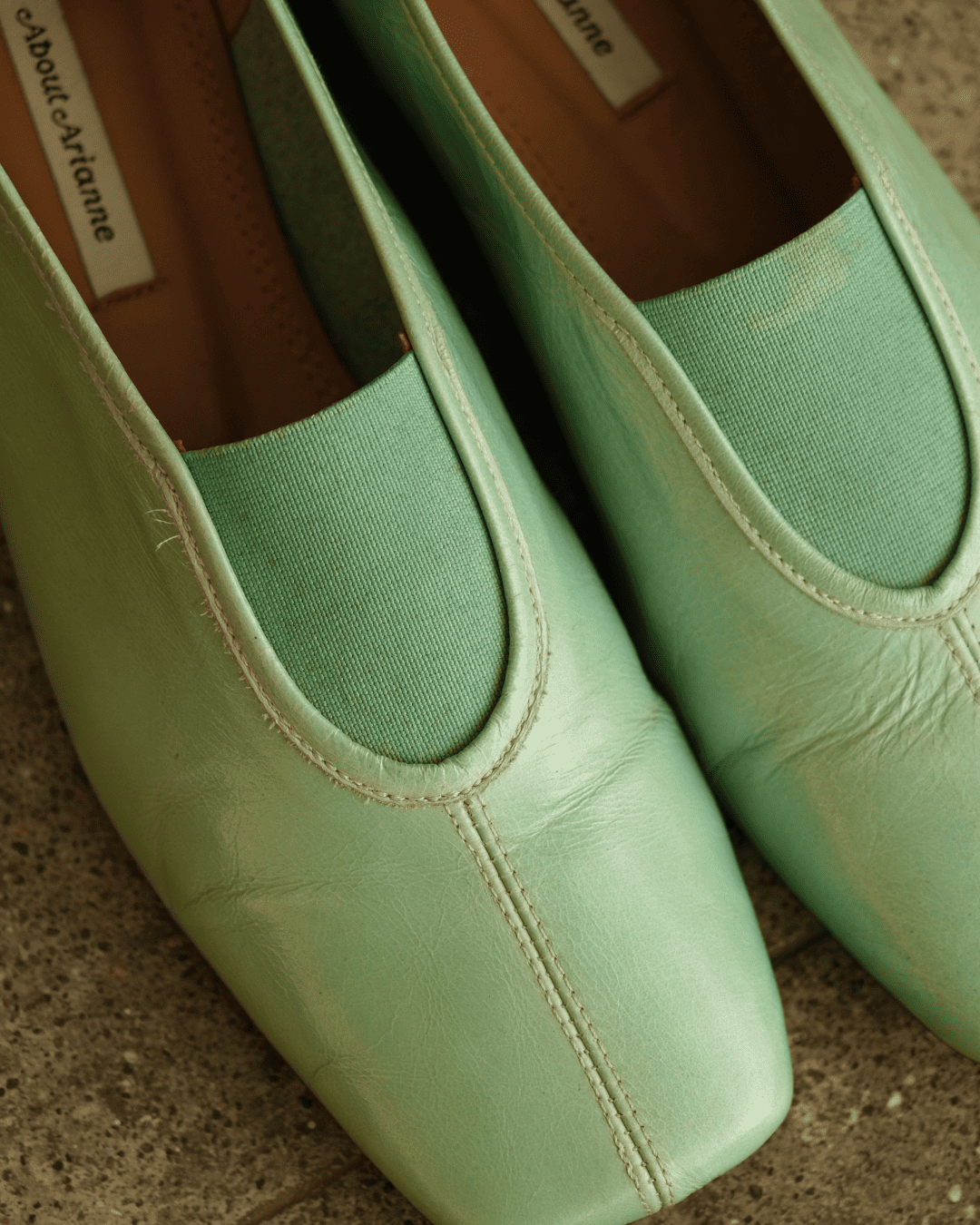 A close-up of mint green slip-on shoes with a textured fabric lining and a tan interior, resting on a textured stone surface. The brand name 'about.