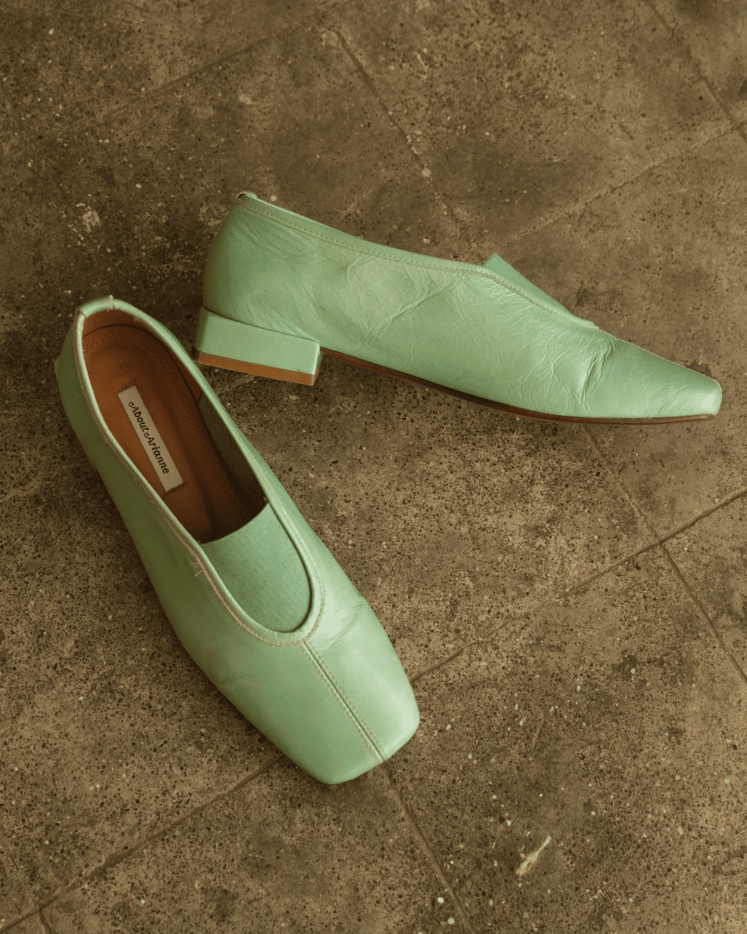 A pair of light mint green leather slip-on shoes with pointed toes and low block heels, resting on a textured brown stone tile floor. The brand name 'SARAGO' is visible on the inner label.