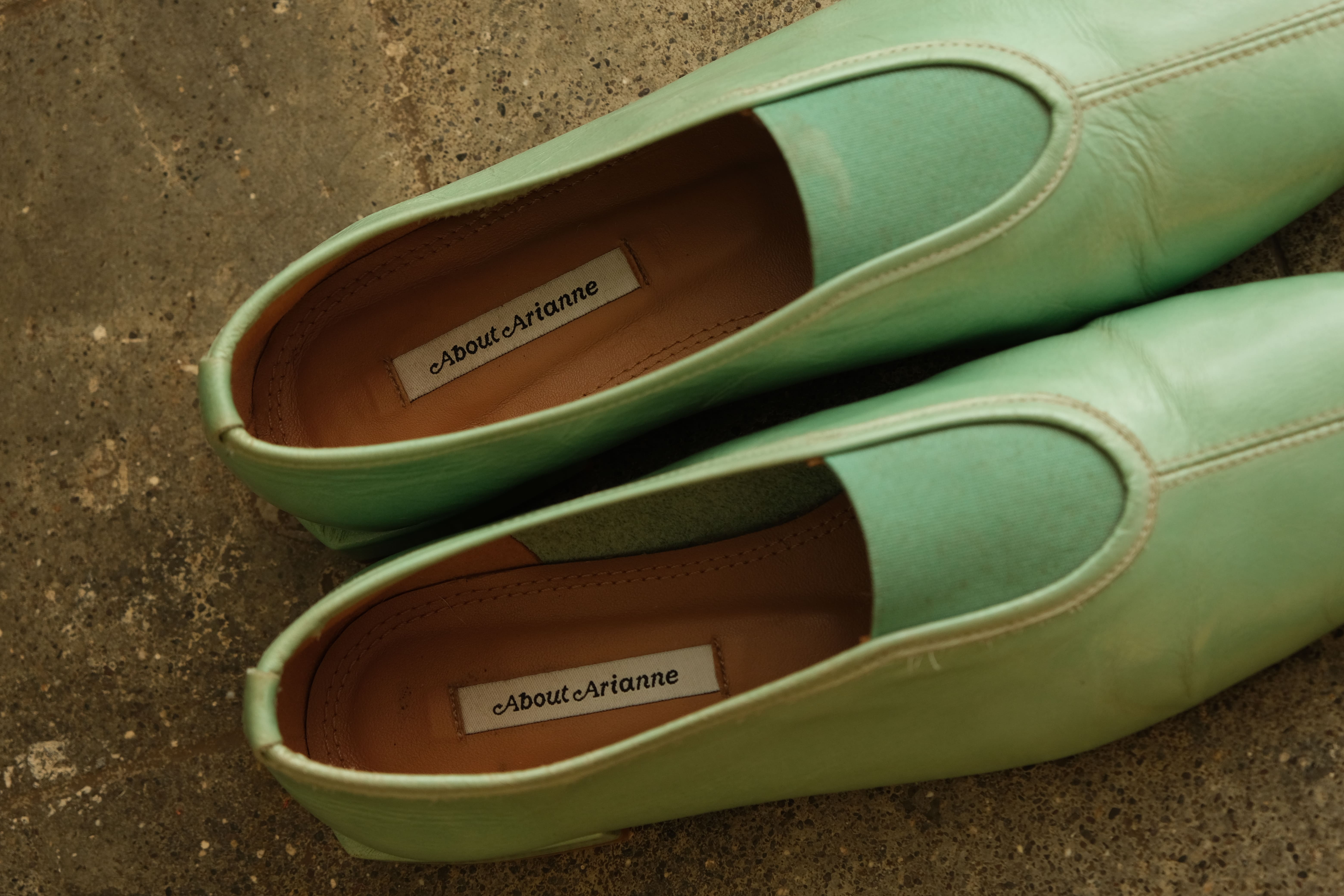 A pair of mint green leather slip-on shoes with pointed toes and a cutout vamp, shown on a textured concrete floor. The insoles display the brand name 'About:Arianne'.