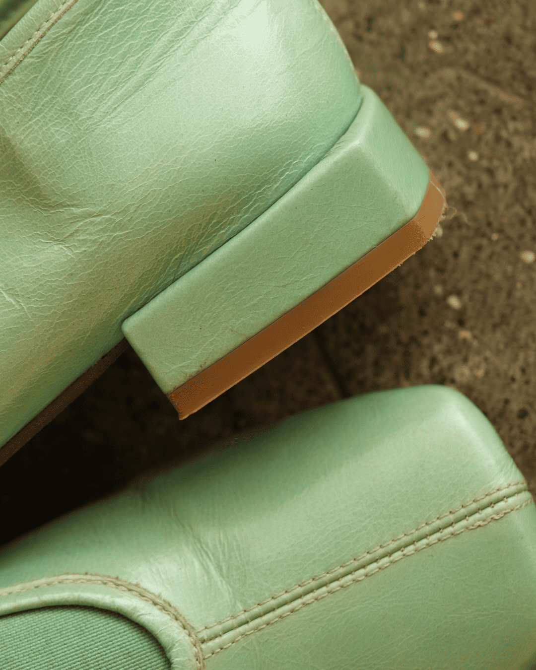 Close-up of mint green leather boots showing the side heel, stitching, and a tan outsole against a textured ground.