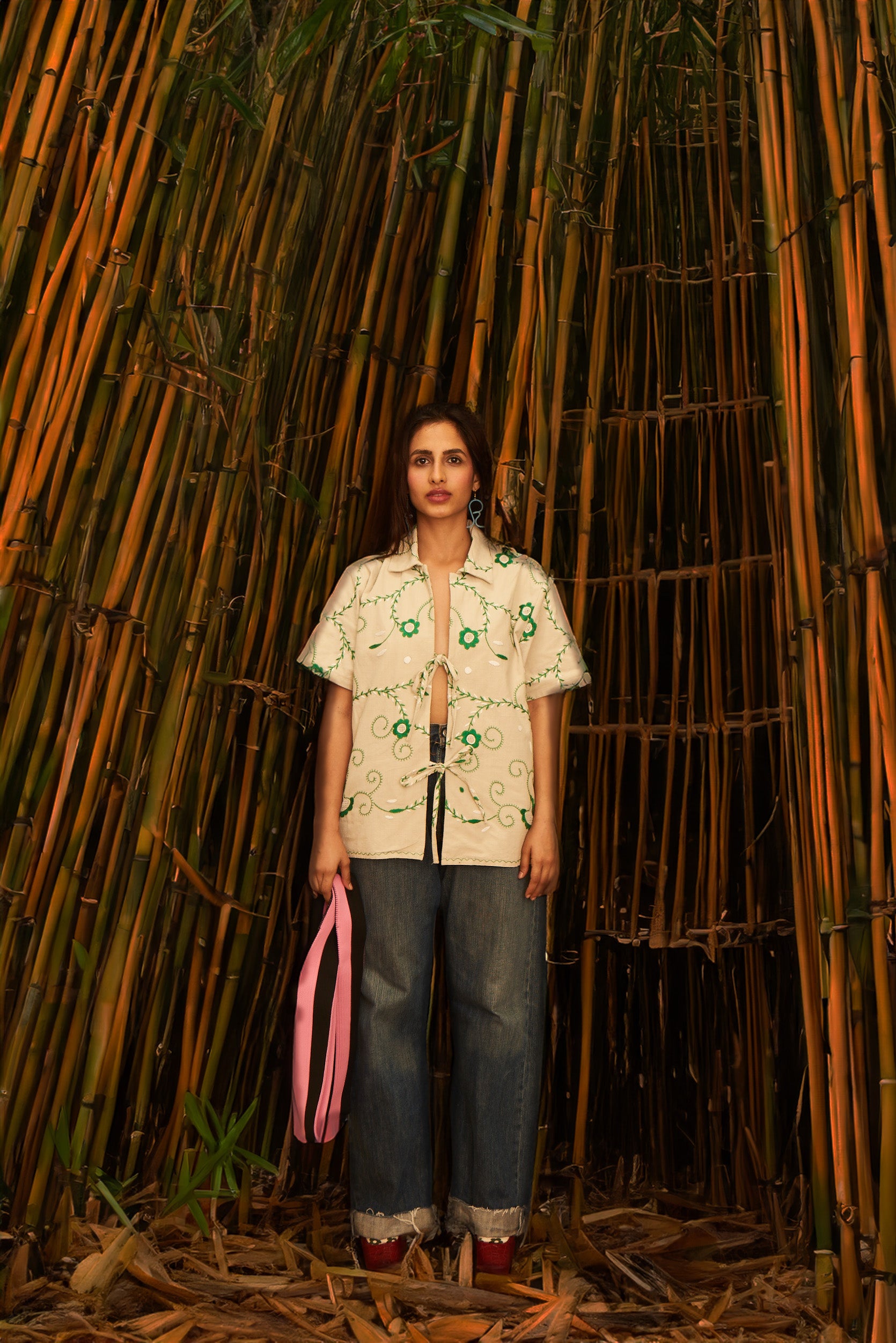 A person standing in a bamboo forest-like setting, wearing a green embroidery shirt with tie-ups for closure, paired with blue jeans and red shoes. Béhen - Green Embroidery Shirt sold by two extra lives