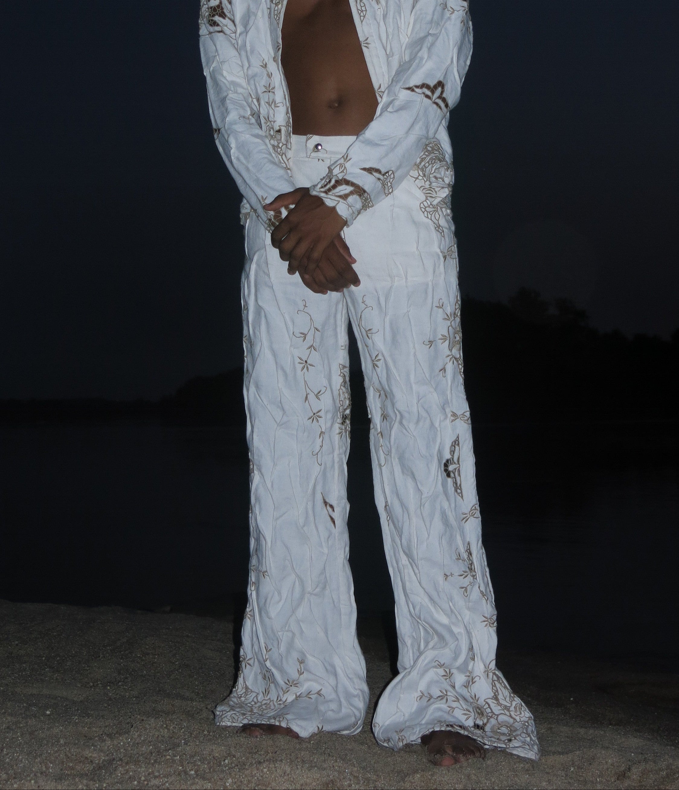 A person standing outdoors at night wearing white pants with a wet/wrinkled effect, resembling a granito texture, typical of traditional madeira embroidery. Béhen - Wet Pants sold by two extra lives