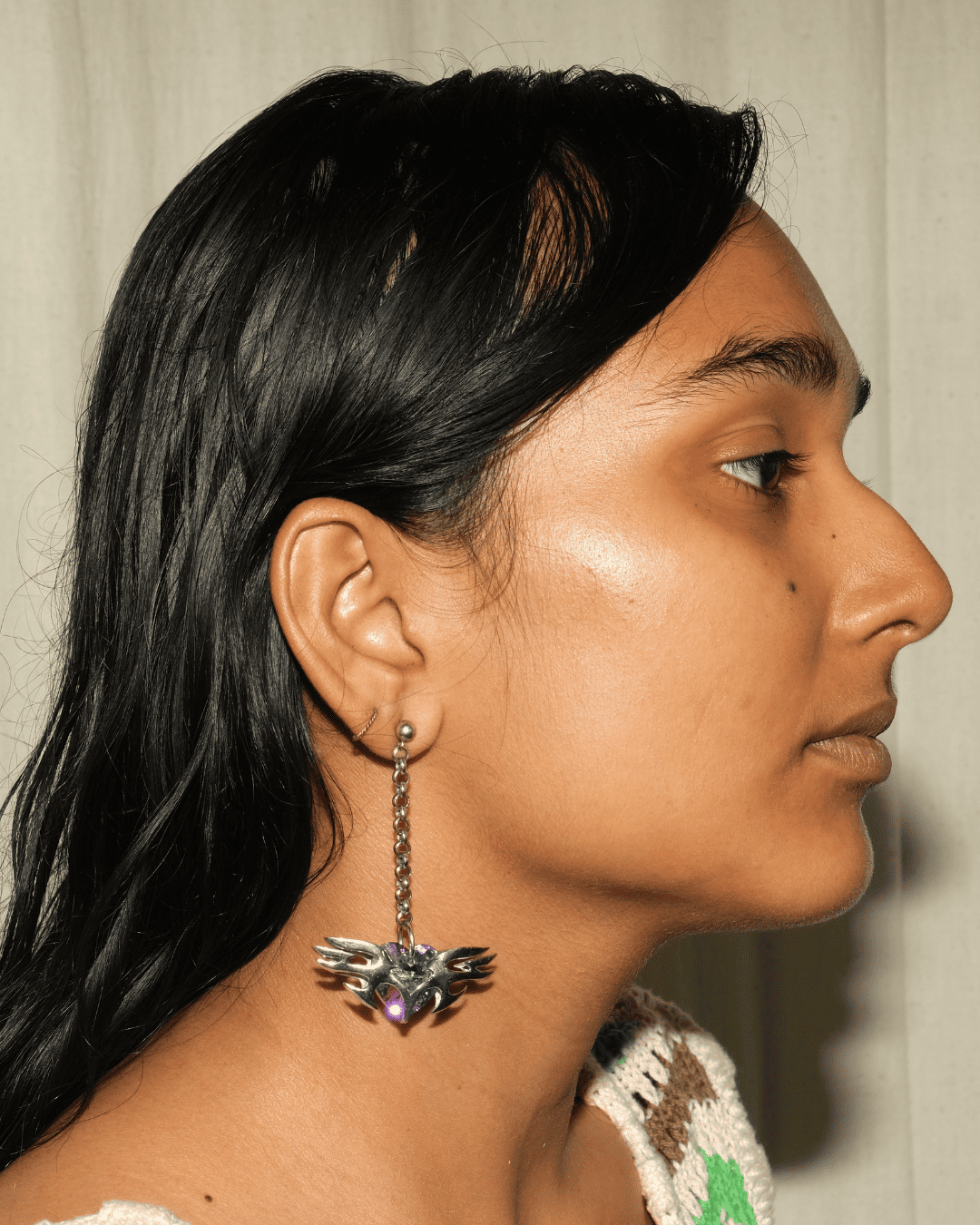 A close-up side profile of a woman wearing a dangling silver earring with a winged emblem and a small purple stone, against a light neutral background.