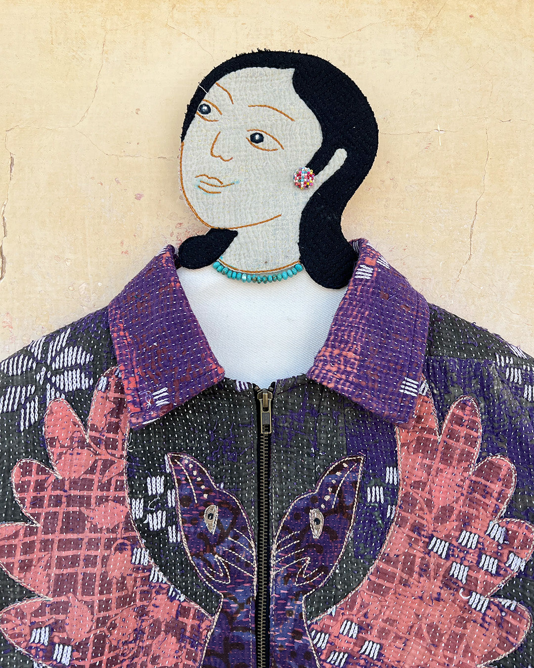 Textile art piece of a person wearing a patterned jacket with floral designs on a beige background DAY-AND-AGE-BHERUNDA-JACKET-TWO-EXTRA-LIVES