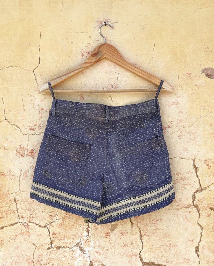 Day and age Blue skorts with white embroidery on a wooden hanger against a textured beige wall. By two extra lives