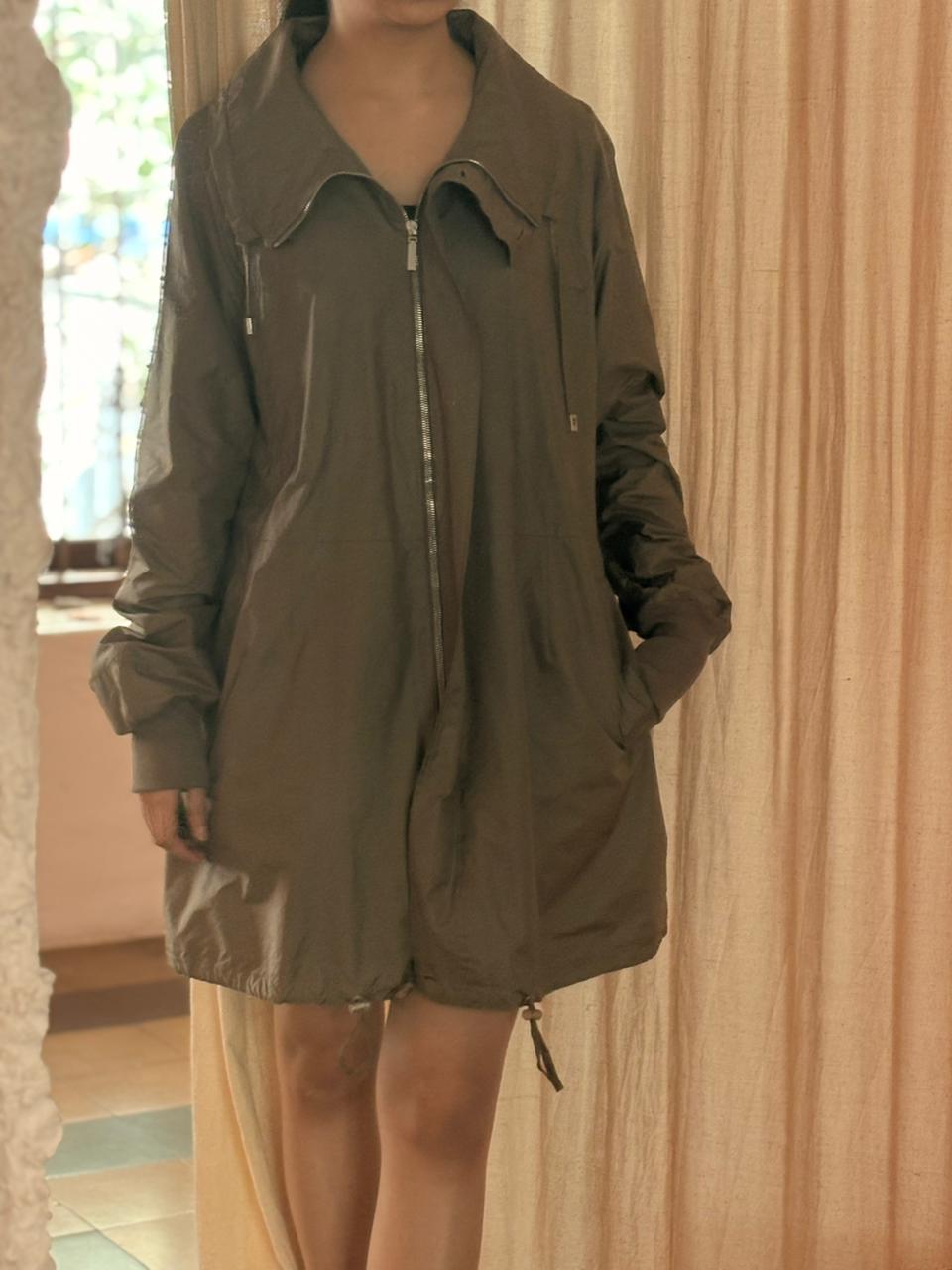 Person wearing a green jacket with a zipper in front of a beige curtain. FROM-THE-MIND-UPCYCLED-VOYAGE-JACKET