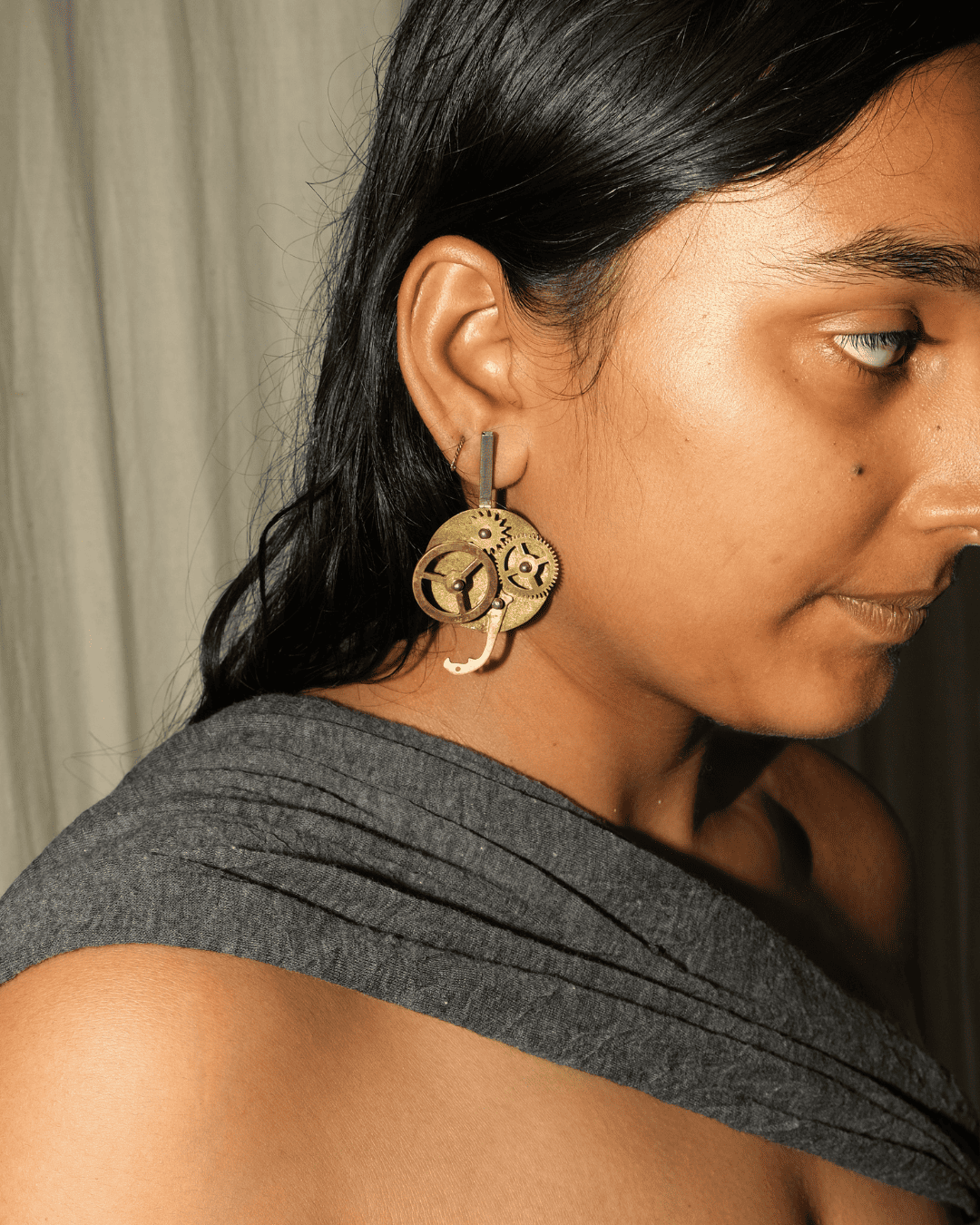 A close-up side profile of a woman wearing a large, intricate gold-toned geometric dangle earring. The earring features layered circular and spiral patterns.