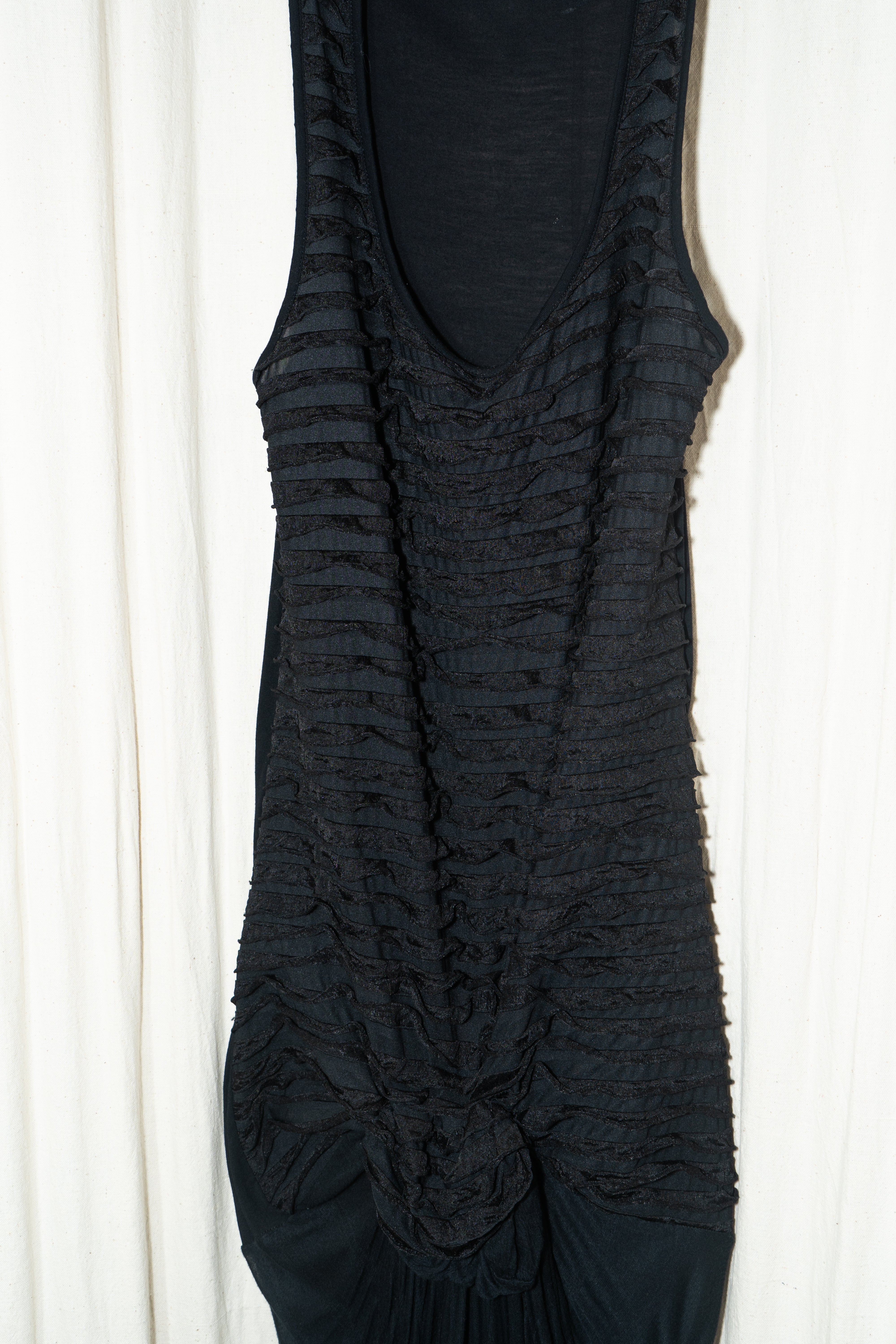 A black maxi gown with flowing trails and gathered fabric at the front, hanging on a hanger against a white curtain backdrop. Kuta House - Trail Gown sold by two extra lives