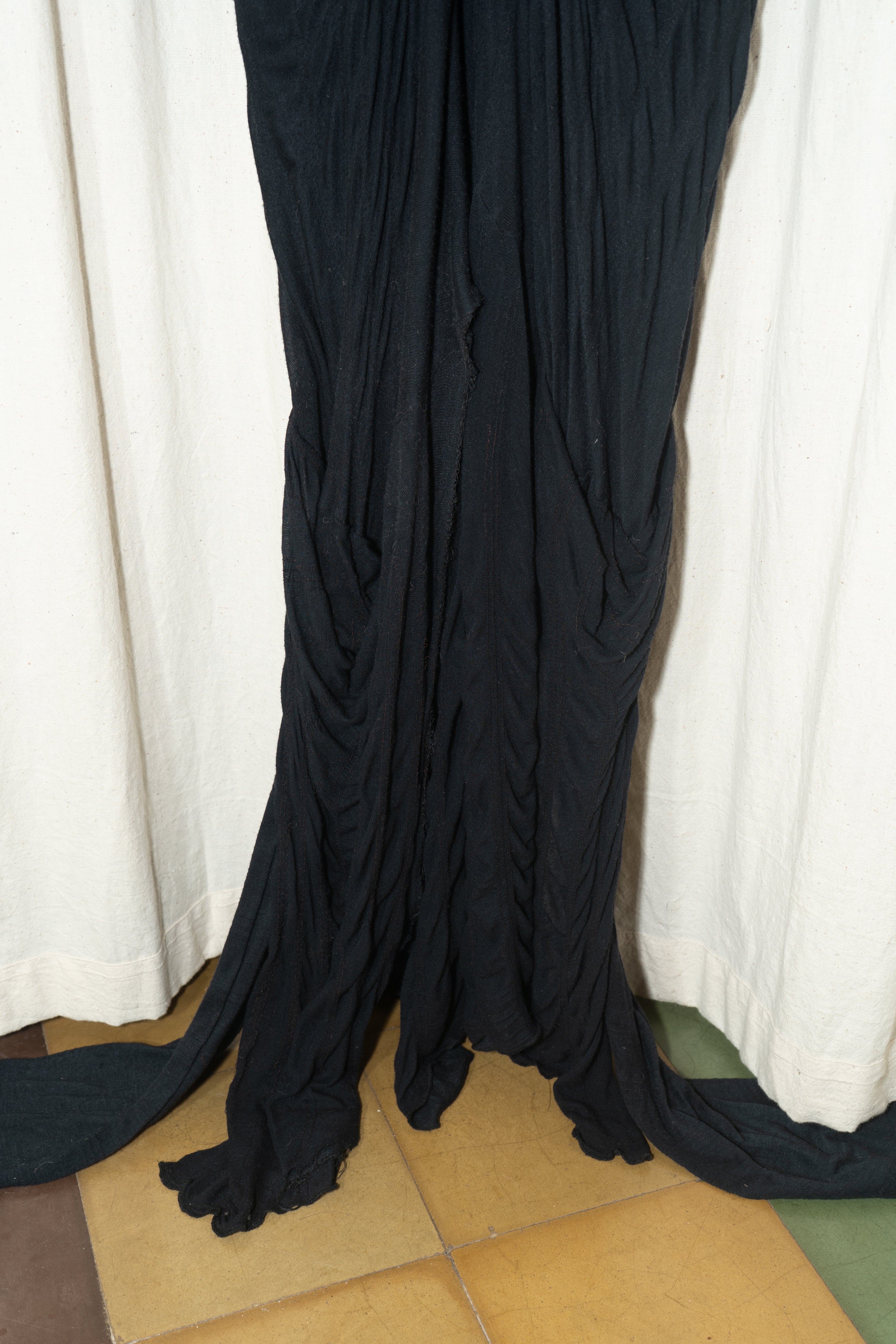 A black maxi gown with flowing trails and gathered fabric at the front, hanging on a hanger against a white curtain backdrop. Kuta House - Trail Gown sold by two extra lives