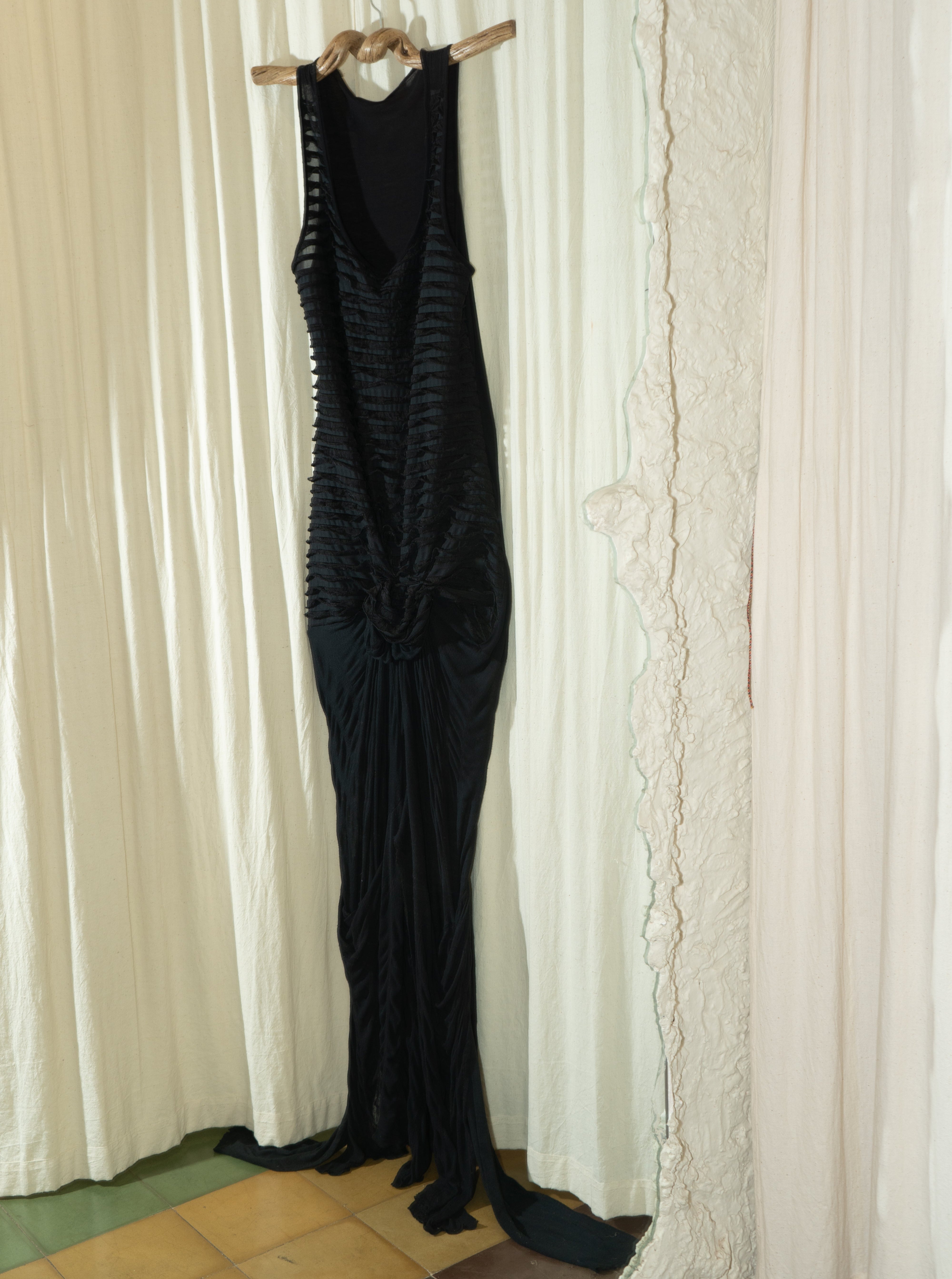 A black maxi gown with flowing trails and gathered fabric at the front, hanging on a hanger against a white curtain backdrop. Kuta House - Trail Gown sold by two extra lives