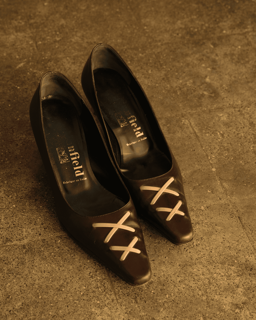 A pair of dark brown pointed-toe pumps with white embroidered X-shaped details on the toes, resting on a textured concrete floor. The brand name 'affliti' is visible on the insoles.