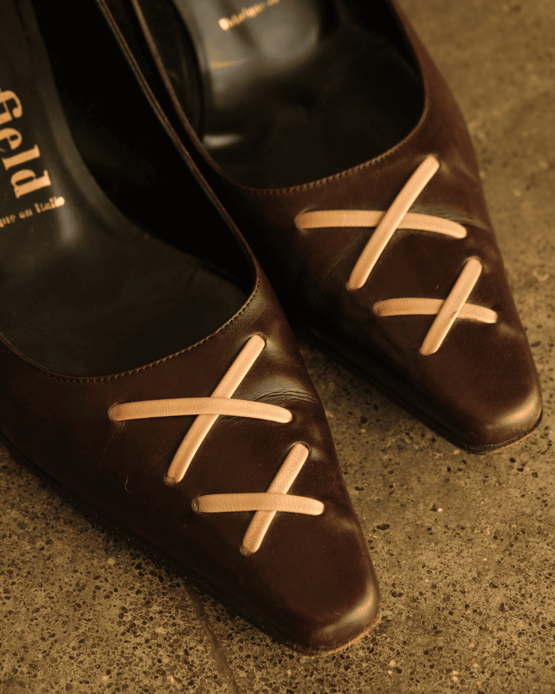 A close-up of a pair of dark brown pointed-toe pumps with light beige faux laces arranged in X shapes across the front, resting on a textured concrete floor. The brand name 'Pleyd' is visible on the insole of the left shoe.