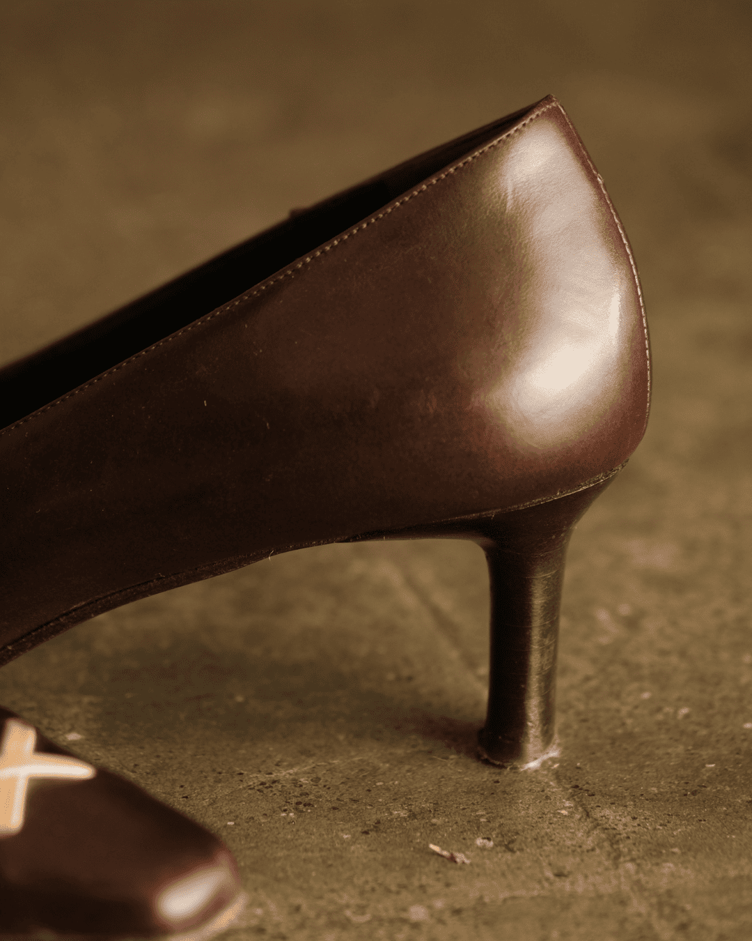 A close-up shot of a dark brown leather high-heeled shoe on a textured, warm-toned surface.