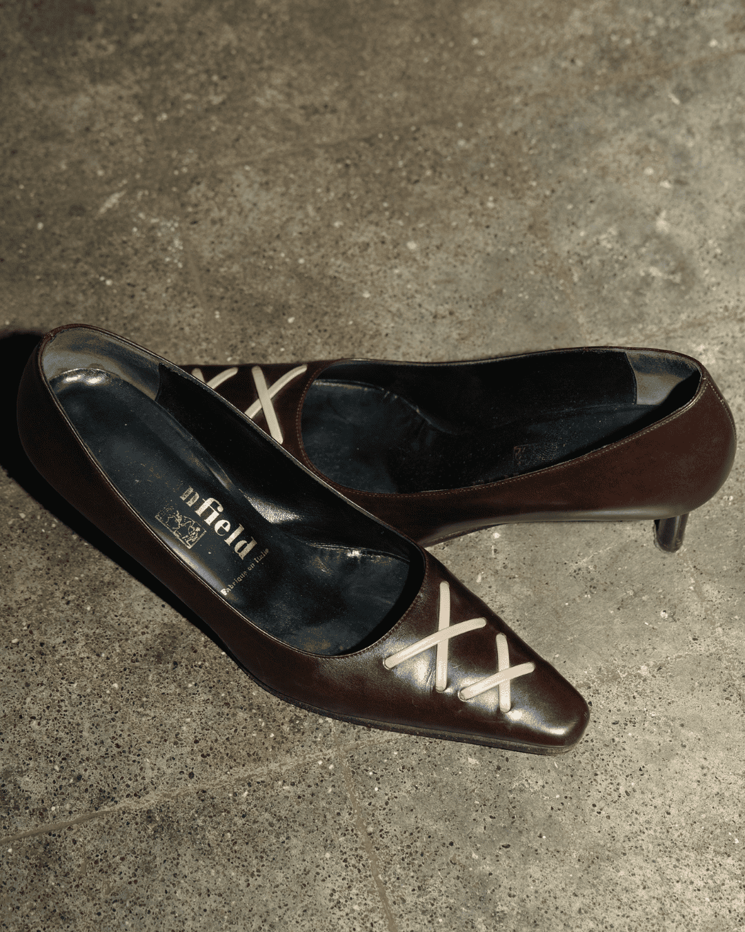 A pair of dark brown pointed-toe pumps with white X-shaped decorative details on the vamp, resting on a textured gray concrete floor. The brand name 'enfield' is visible on the insole.