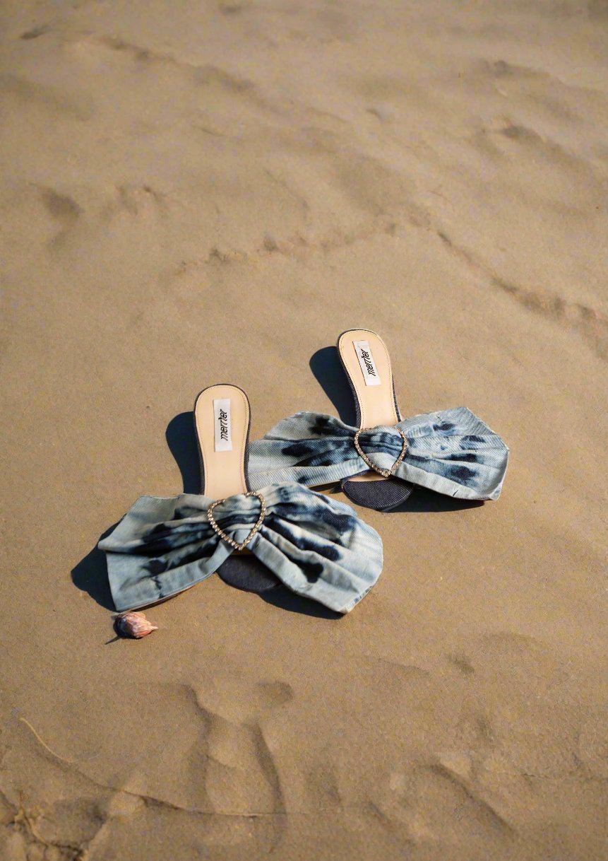 A pair of denim blue heels with oversized bows and sparkling rhinestone hearts at the center, placed on a textured background. Merrfer - Bow Denim Heels sold by two extra lives