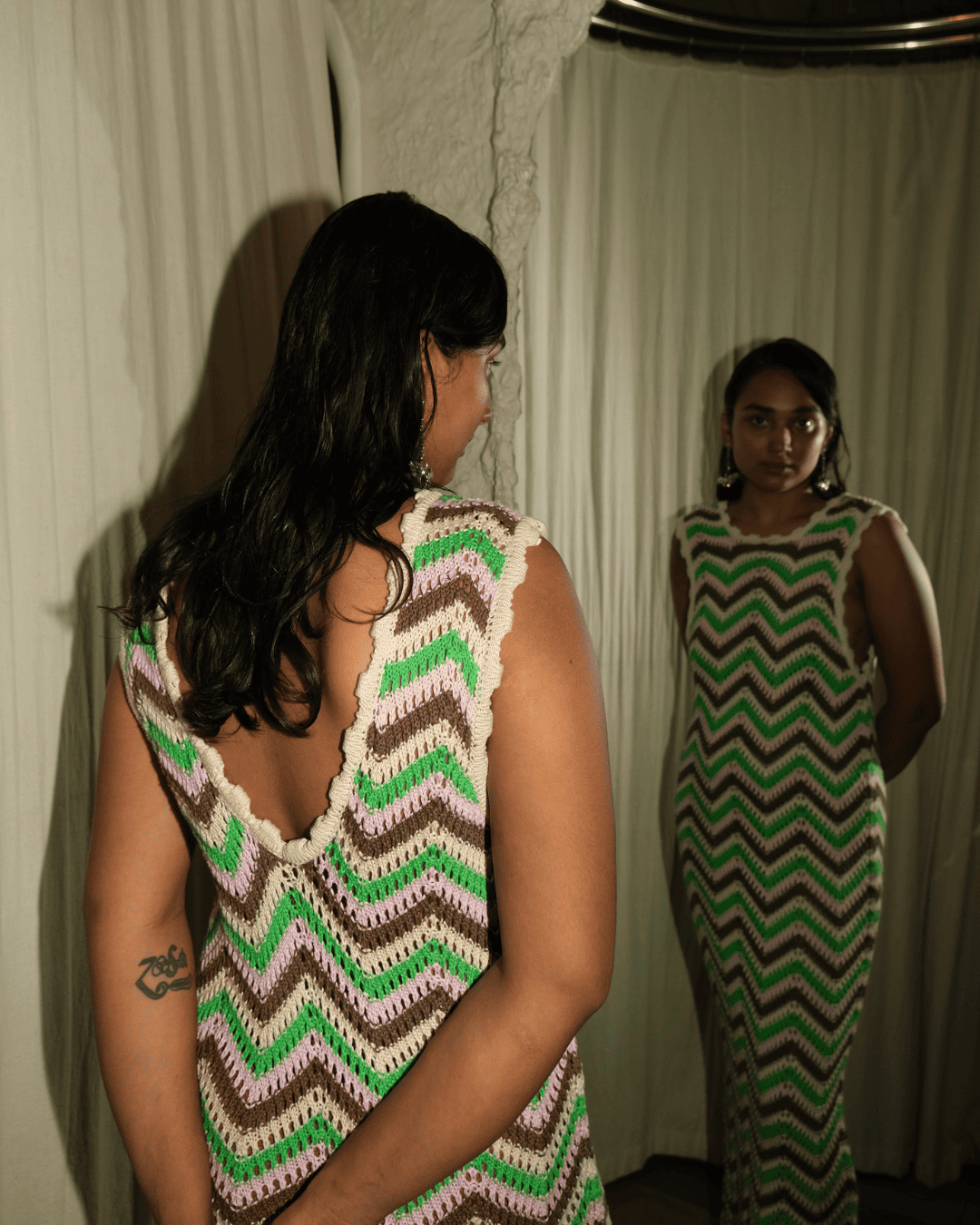 A woman stands with her back to the camera wearing a sleeveless crocheted dress with a green, brown, and white chevron pattern and an open back. She is standing in front of a mirror that reflects her front view of the dress.