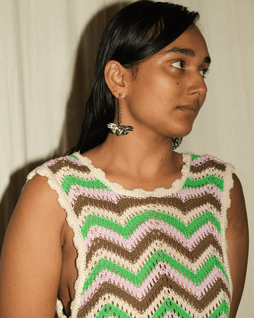A woman wearing a sleeveless crocheted top with a colorful chevron pattern in green, brown, light pink, and cream, paired with silver butterfly earrings, set against a neutral draped fabric background.