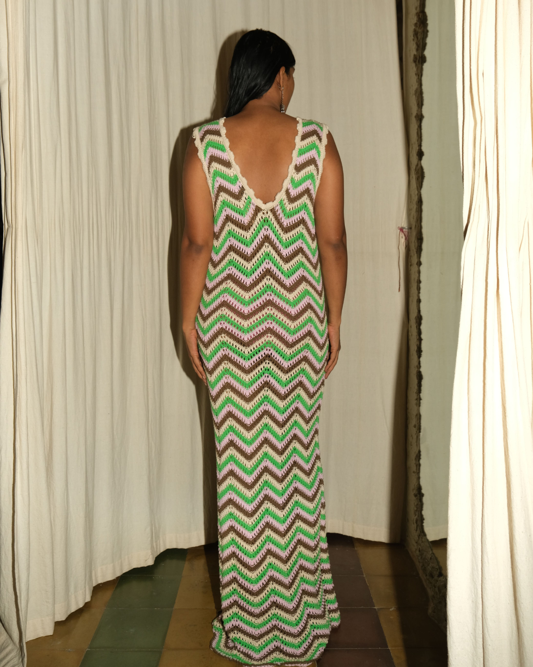 A woman viewed from behind wearing a sleeveless, floor-length knit dress with a green, brown, and white chevron pattern, standing in front of white curtains.