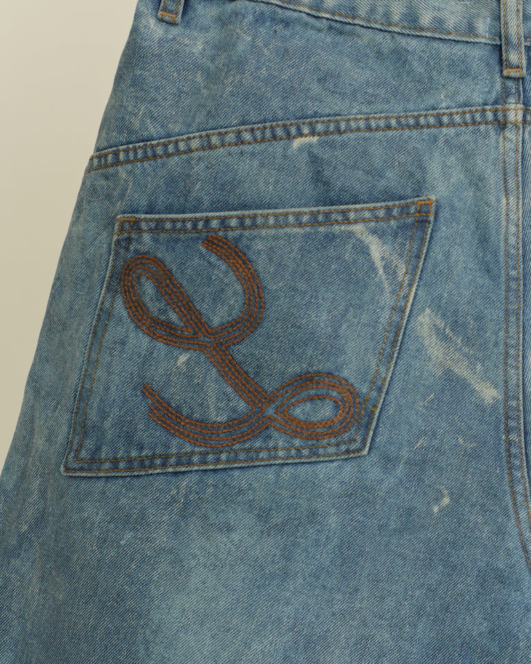 Close-up of a blue denim pocket with a brown embroidered letter 'L' on a beige background