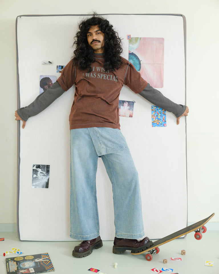 Person wearing a brown t-shirt with text and blue jeans standing next to a large white frame on a white wall.