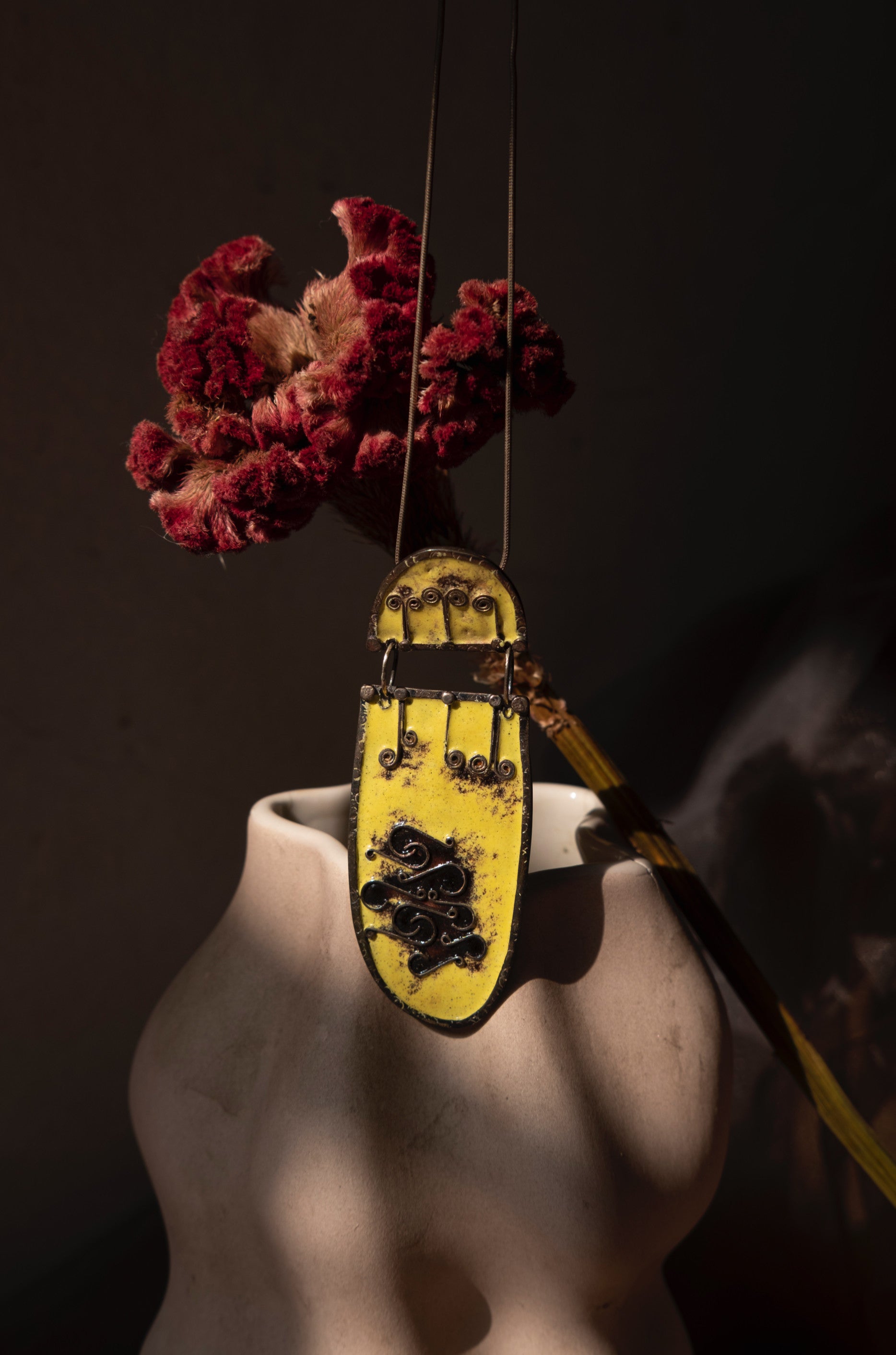 A metallic locket neckpiece with a yellow and black abstract design, displayed on a mannequin bust, with a red flower attached to it. Peca-Joyeria-Poetica-Metallic-Locket-Neckpiece sold by two extra lives