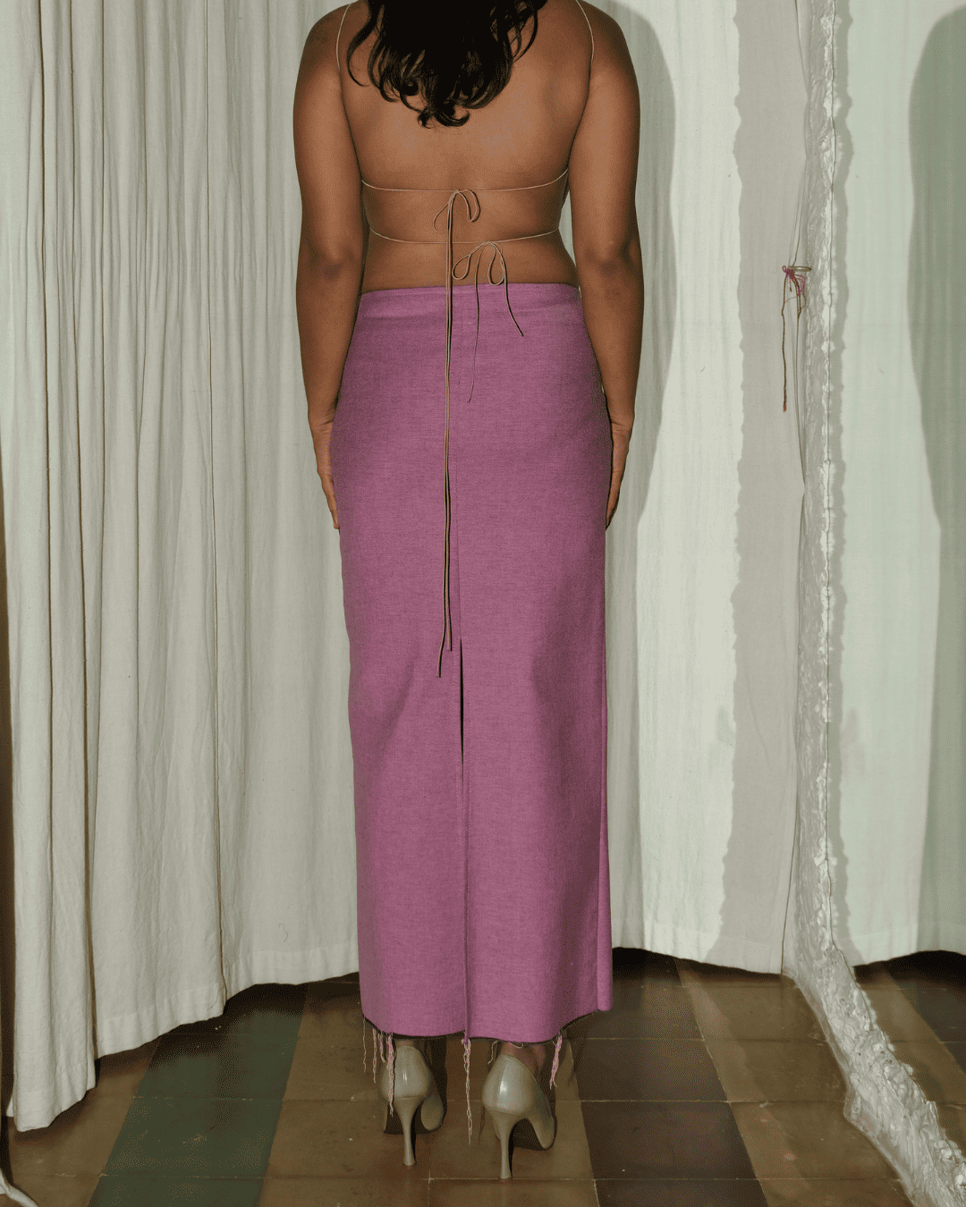 A woman shown from the back wearing a dusty pink knit midi skirt with a front zipper and tie detail at the waist, paired with beige high heels, standing in front of white curtains.