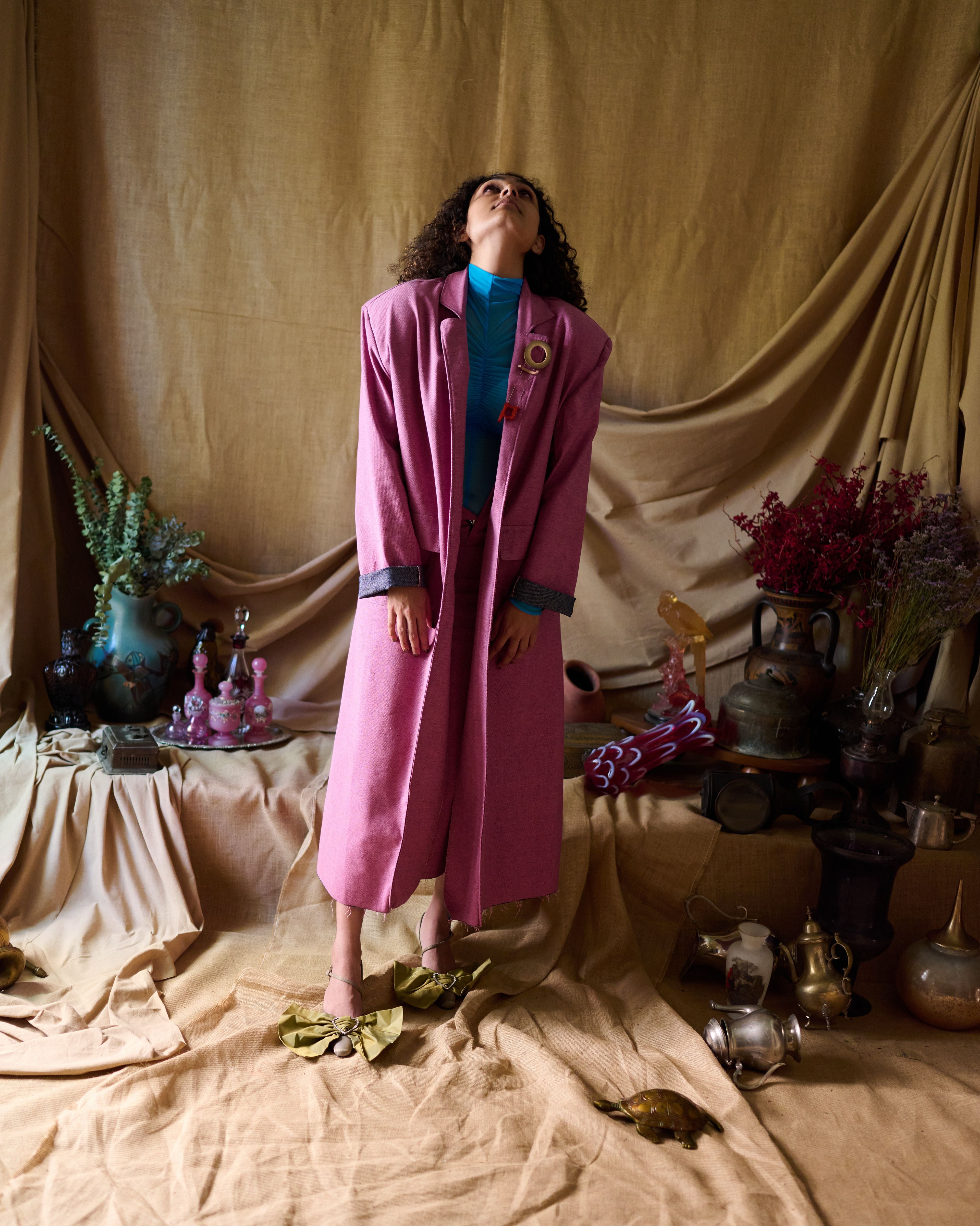 A person wearing a pink denim trench coat with a tailored fit, standing in a room with a beige background, surrounded by various decorative items. Roa-Round-the-World-Pink-Trench-Coat-two-extra-lives