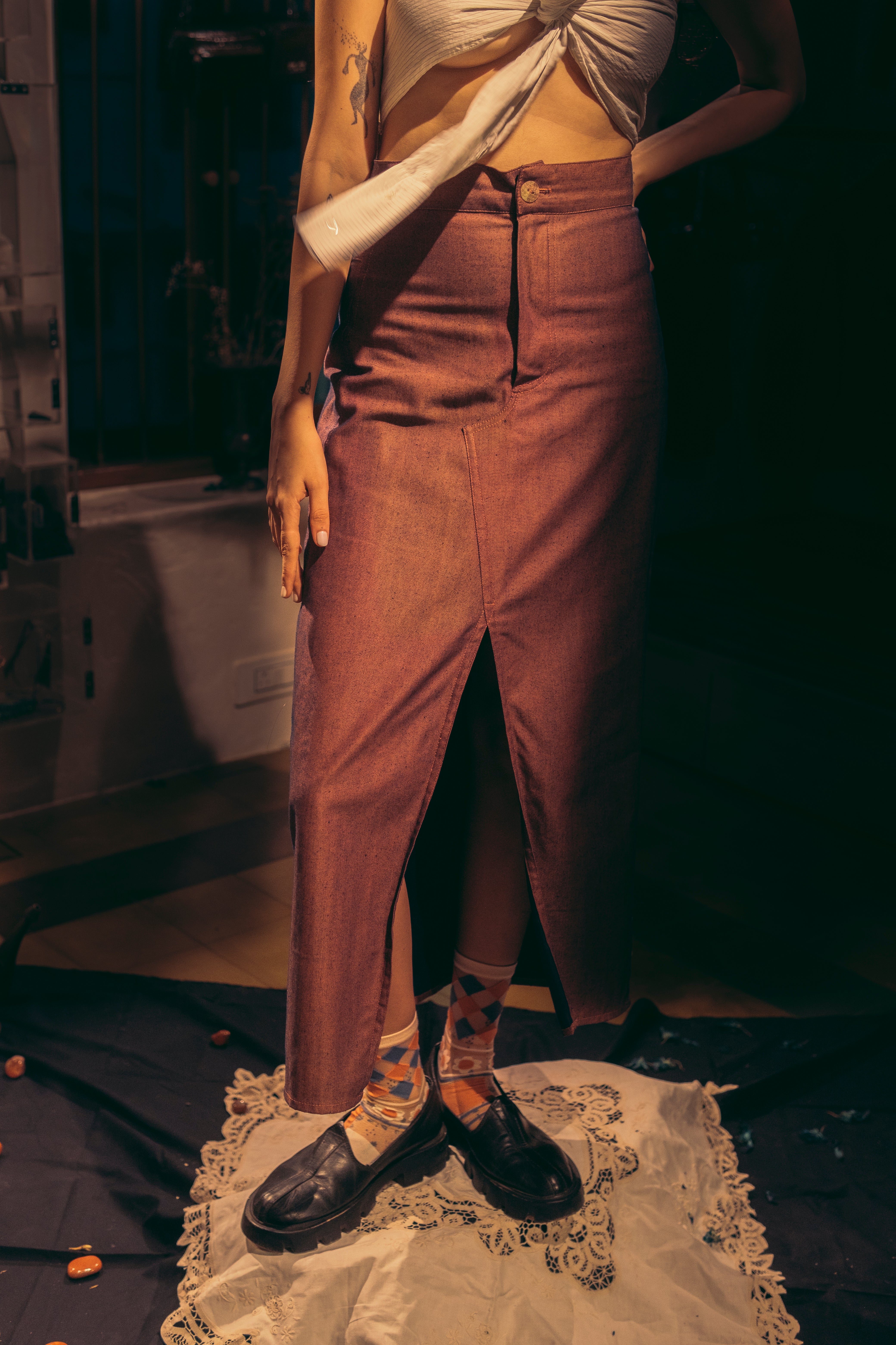 A person wearing a deep wine-colored denim skirt with a front slit, paired with a white cropped tank top and red-brown sandals, standing in a room with a reflective floor. Roa-Wine-Slit-Skirt-two-extra-lives