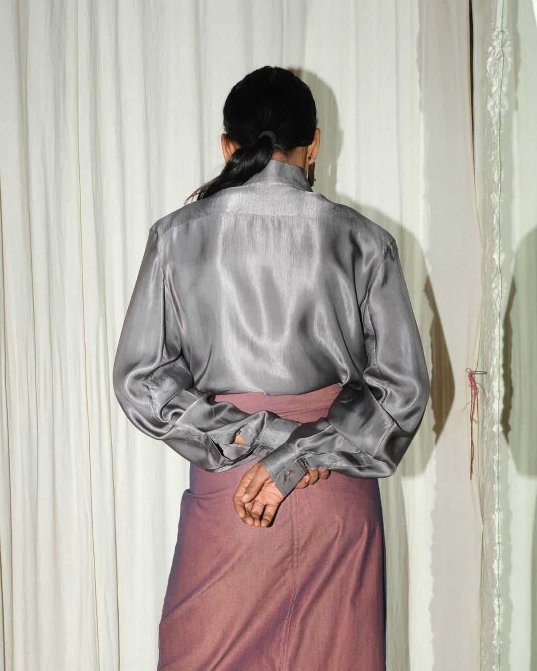 A person shown from the back wearing a shiny gray long-sleeve top and a dusty rose skirt, standing in front of a white curtain.