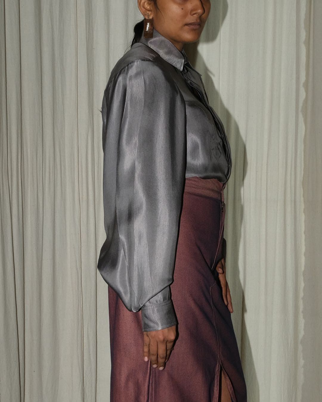 A model shown from the side wearing a shiny dark gray satin long-sleeve blouse and a dark purple skirt, standing in front of a draped off-white curtain.