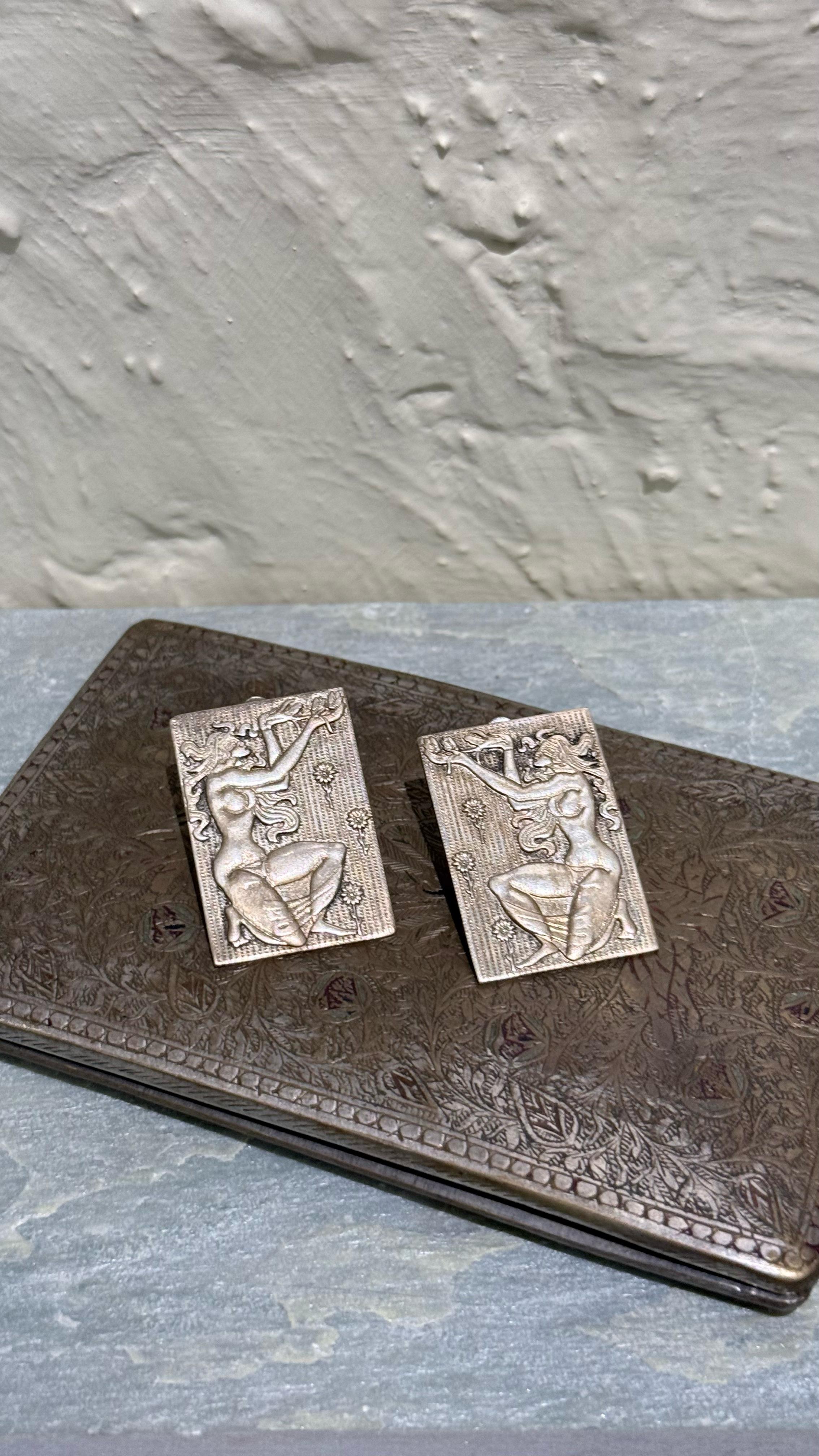 A pair of silver clip-on earrings with embossed tribal depictions presented on a textured surface.