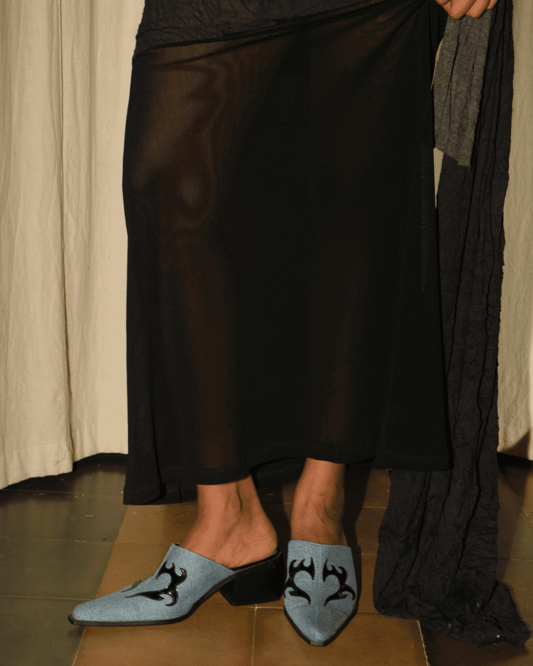 A person wearing a sheer black maxi skirt and light blue pointed-toe mule heels with black embroidered wing-like motifs, standing indoors against a draped curtain backdrop.