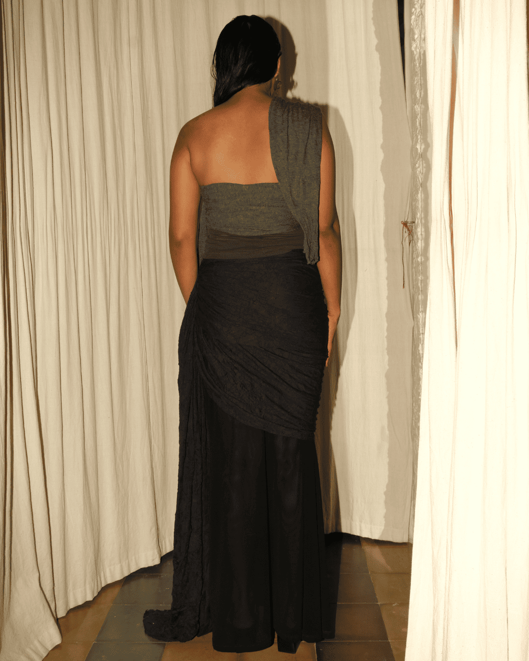 A woman viewed from behind wearing a dark gray strapless top with a draped shawl and a long black draped skirt, standing in front of cream-colored curtains.