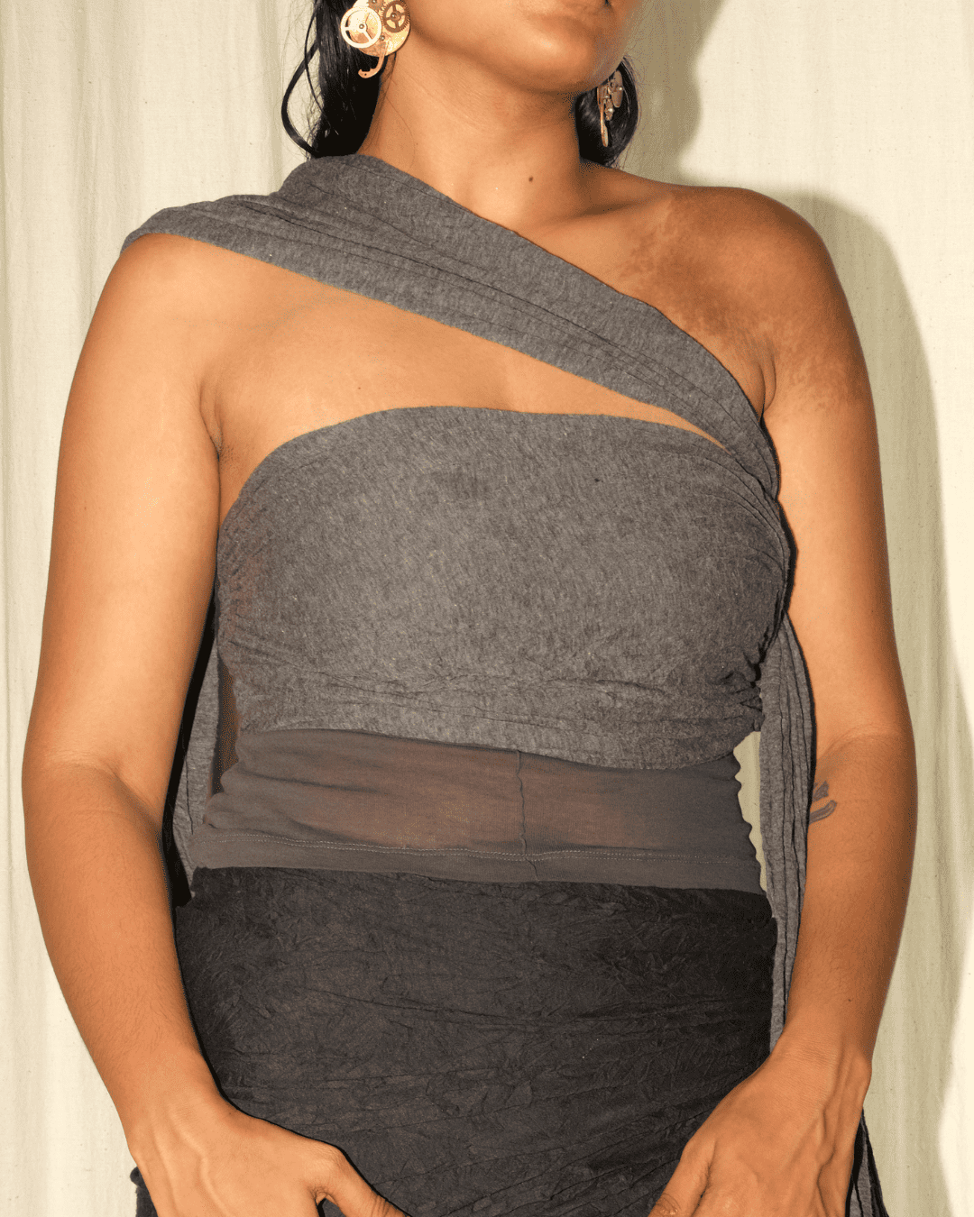 A woman wearing a dark gray one-shoulder wrap top with a sheer mesh band at the waist, paired with a dark skirt, against a light curtain background.