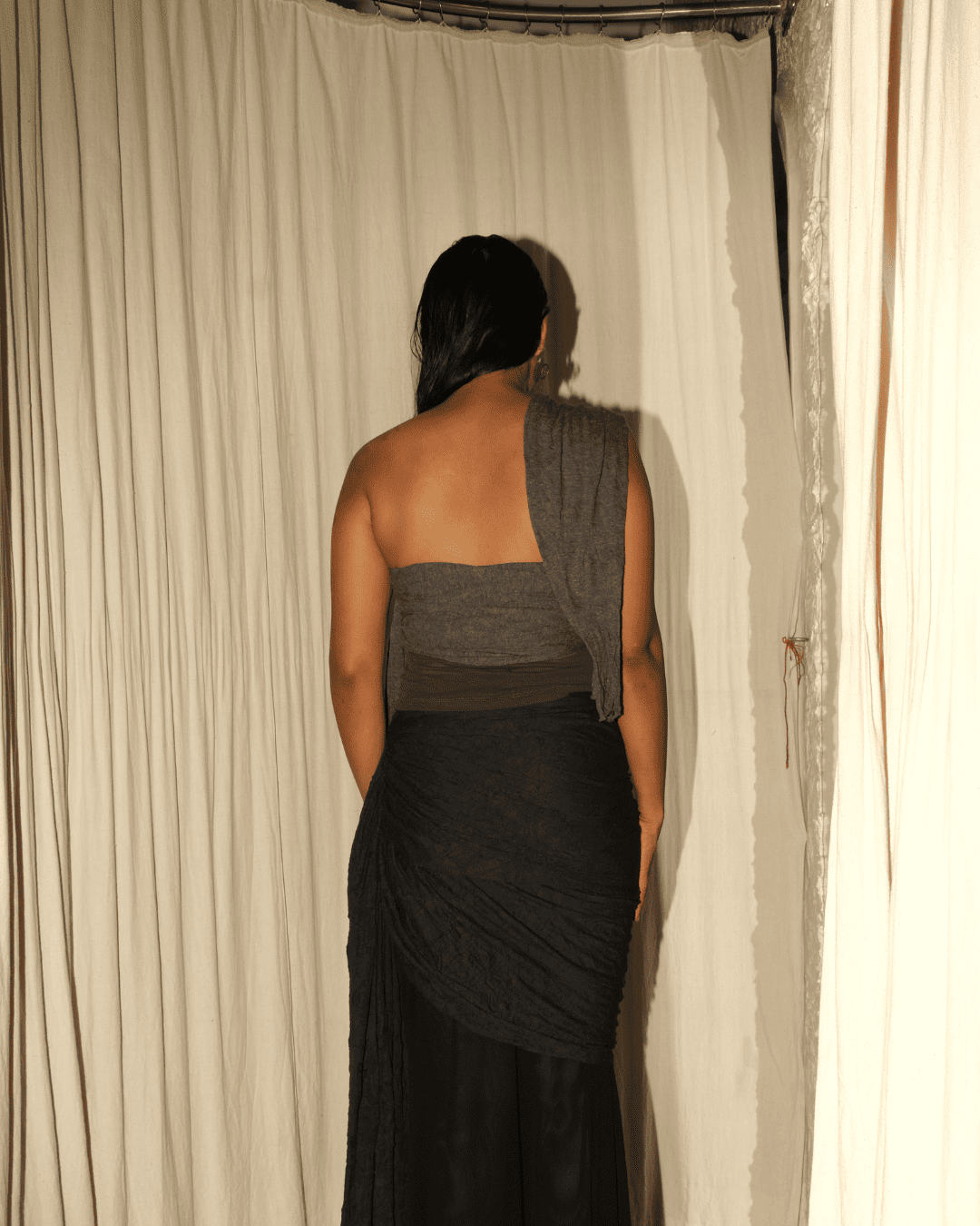 A woman viewed from behind wearing a dark gray strapless bandeau top with a draped shawl over one shoulder and a long dark skirt, standing in front of cream-colored curtains.