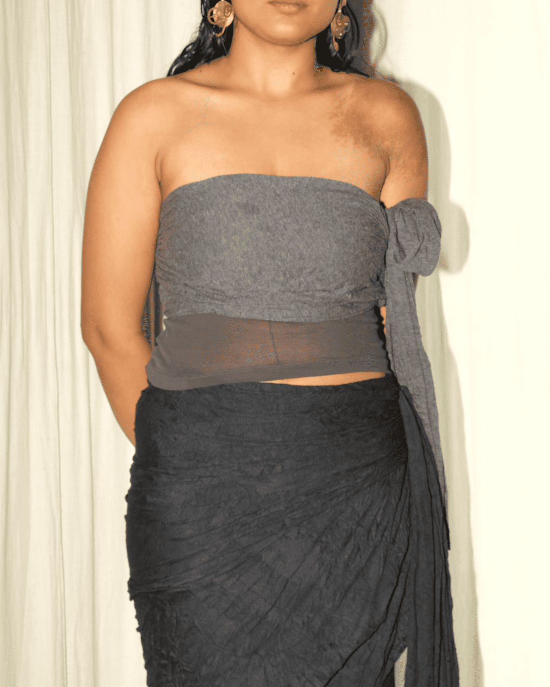 A woman wearing a strapless gray crop top with a sheer mesh band and a matching dark gray draped wrap skirt, shown against a light curtain background.