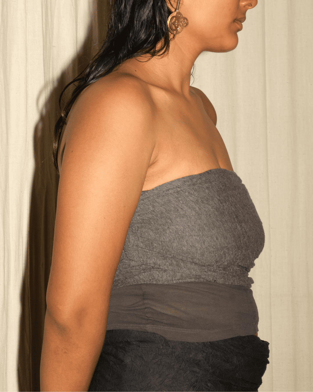 A woman shown in side profile wearing a strapless gray bandeau-style top with a sheer dark gray waistband and a black textured bottom, standing against a light curtain background.