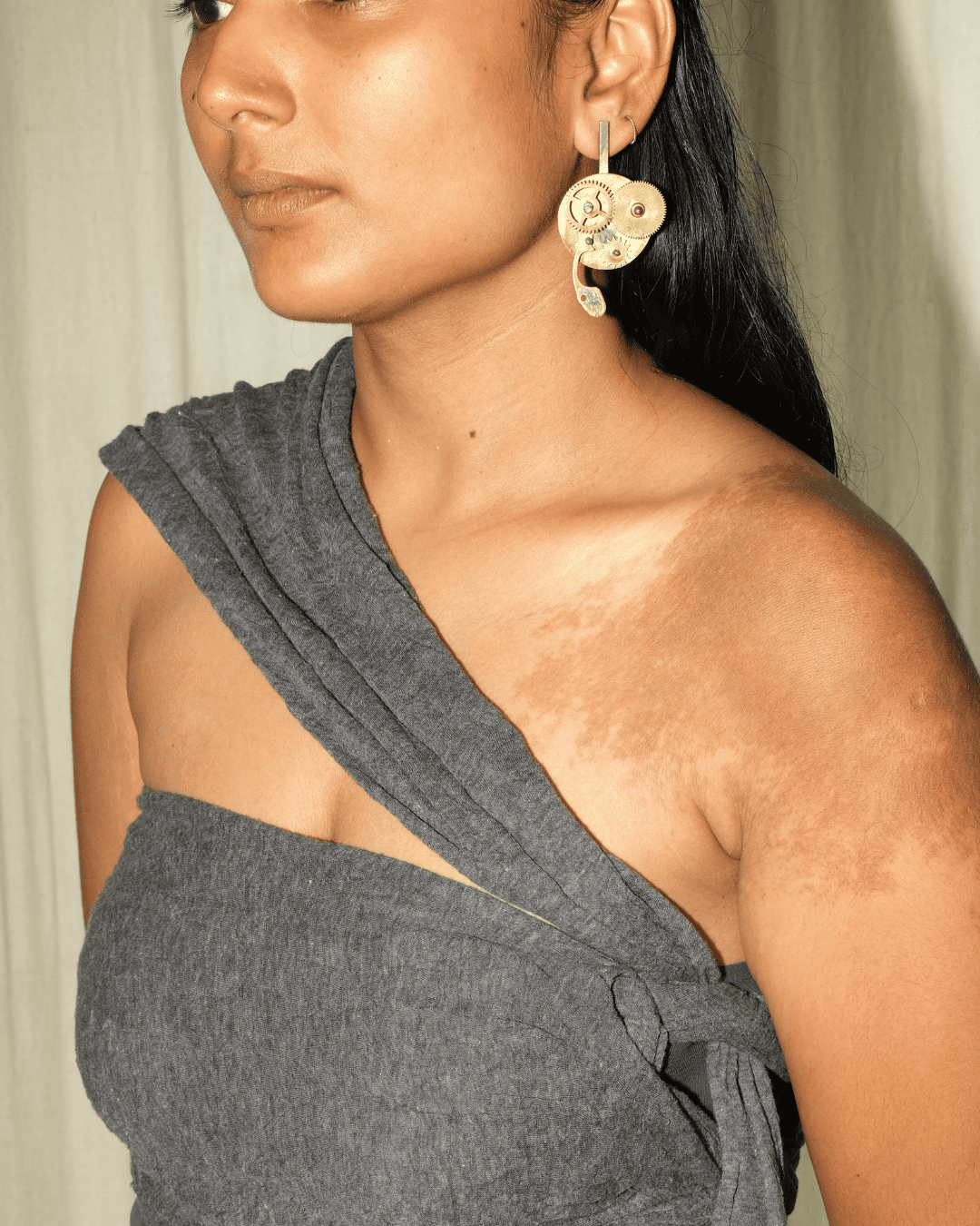 A close-up of a woman wearing a dark gray draped one-shoulder top and a large gold floral drop earring.
