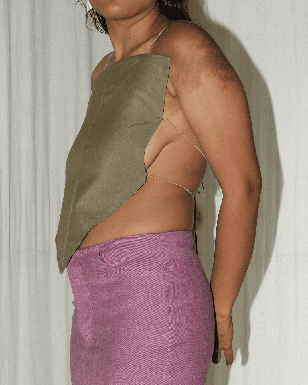 A side-profile view of a model wearing a one-shoulder olive green top with thin spaghetti straps and a purple skirt, standing against a white curtain background.