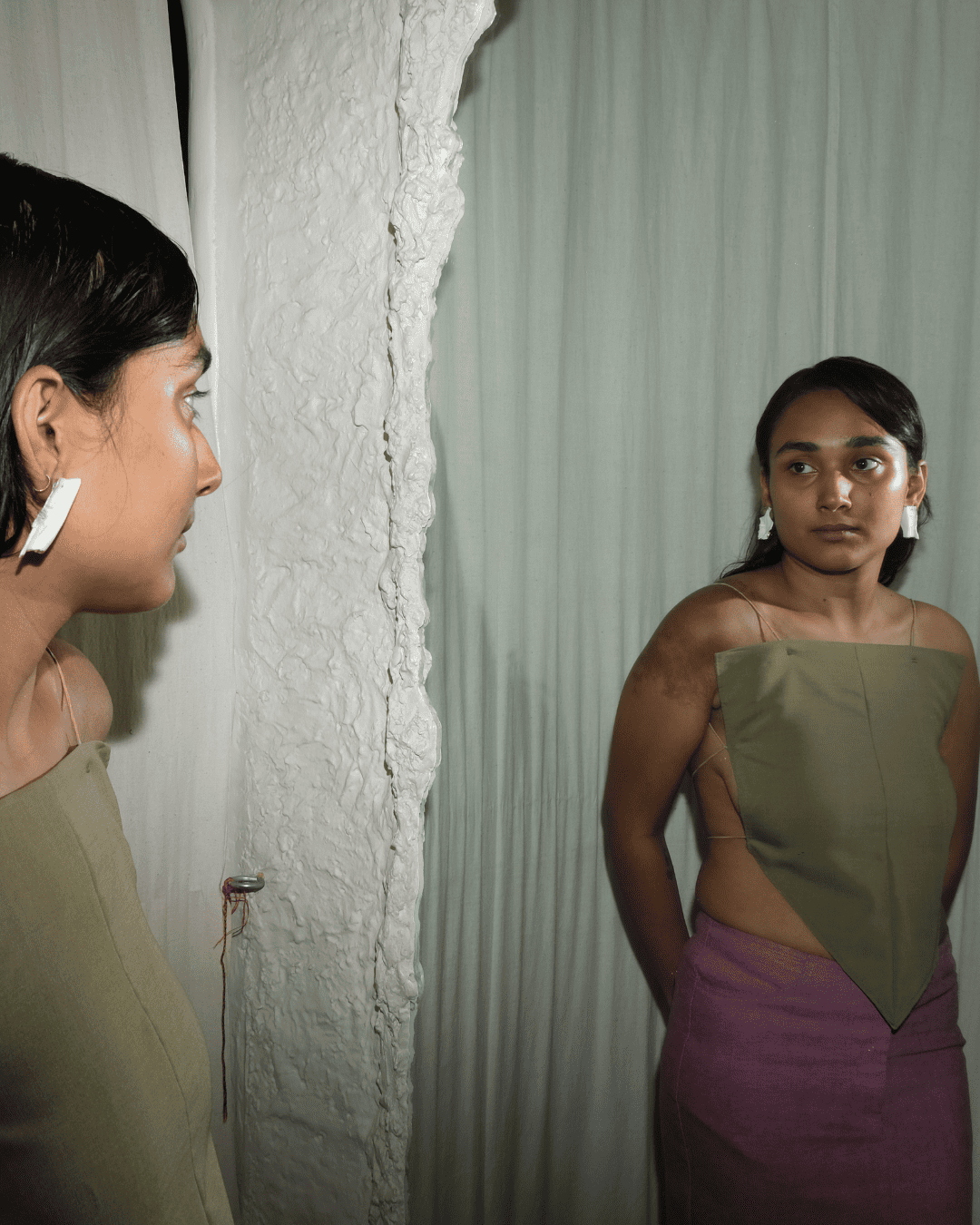 A woman wearing a sleeveless olive green top and purple skirt stands in front of a textured white wall, looking at her reflection in a mirror. She is wearing white rectangular earrings.