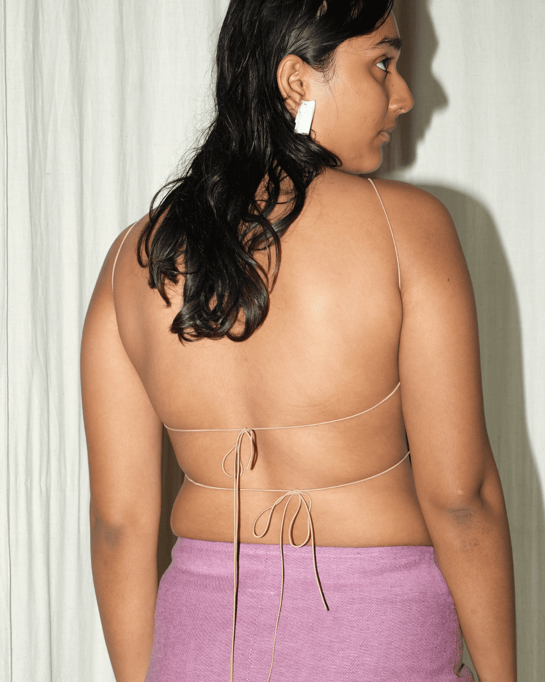 A woman shown from the back wearing a light beige string bikini top with thin straps and a tie-back design, paired with a lavender skirt. She is standing against a white curtain background.
