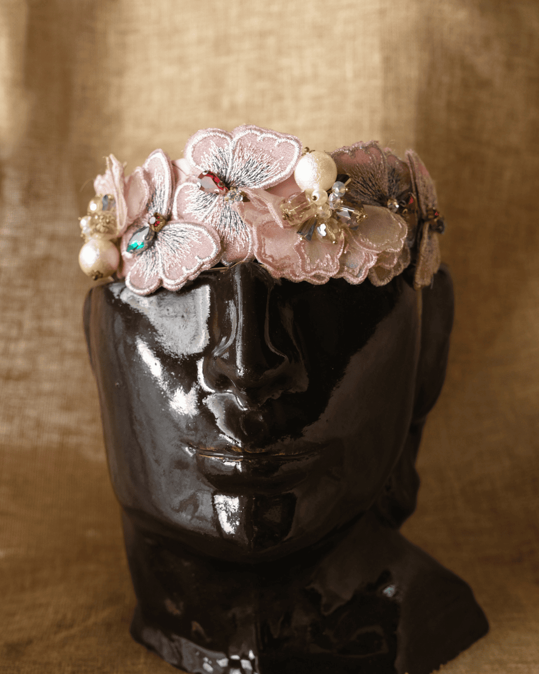 A floral headband with embroidered pink and beige flowers, pearl-like beads, and rhinestone accents, displayed on a glossy black sculptural head against a warm tan fabric background.