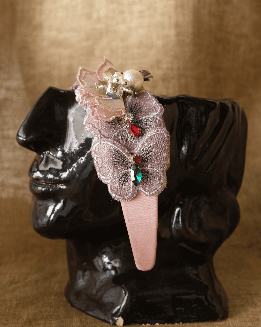 A pink plastic hair claw clip decorated with a light pink lace butterfly, a pearl bead, and small metallic charms, displayed on a glossy black head-shaped stand against a textured tan fabric background.