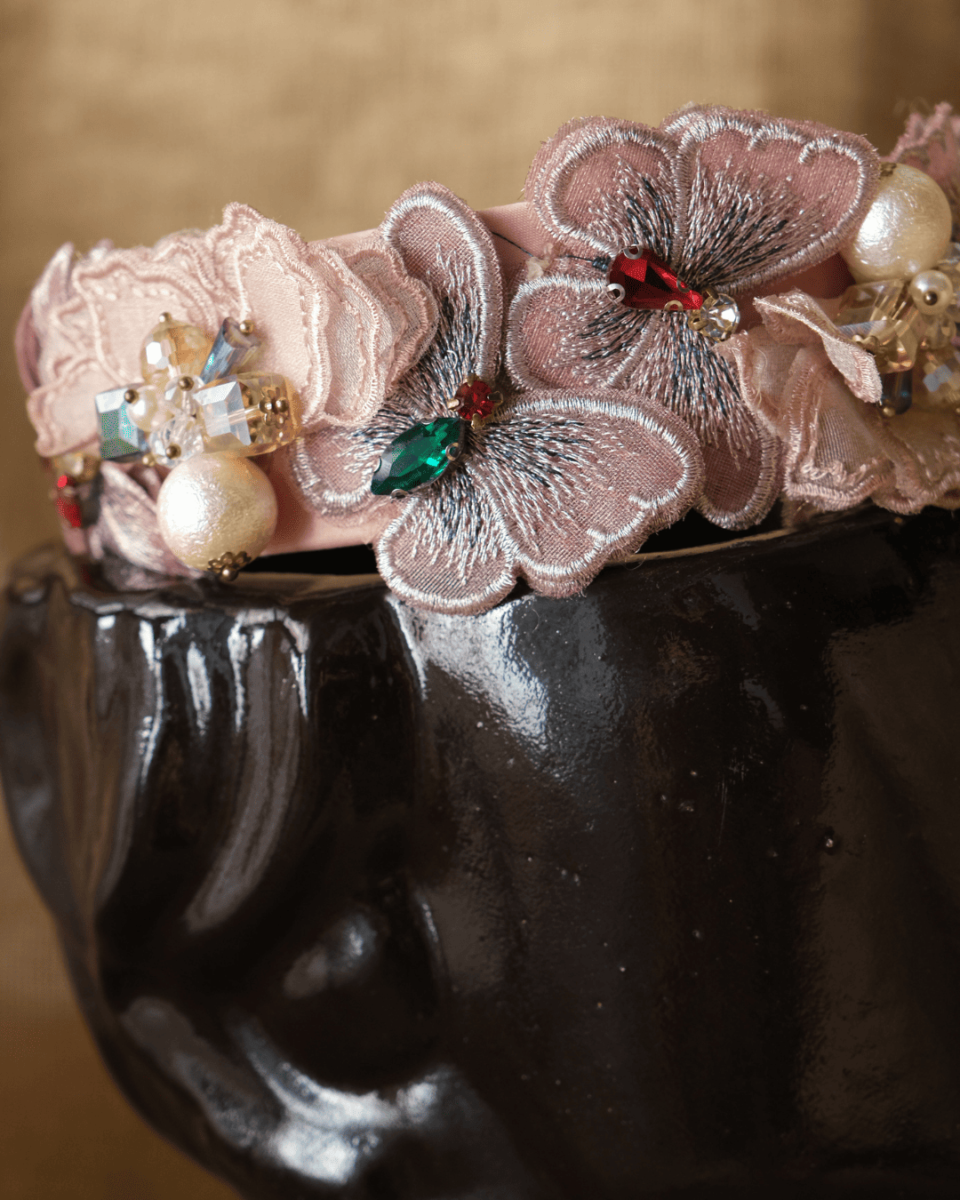 A close-up of a decorative floral headband with layered pink fabric flowers, pearl-like beads, and small jewel embellishments, displayed on a glossy black bust against a warm beige background.
