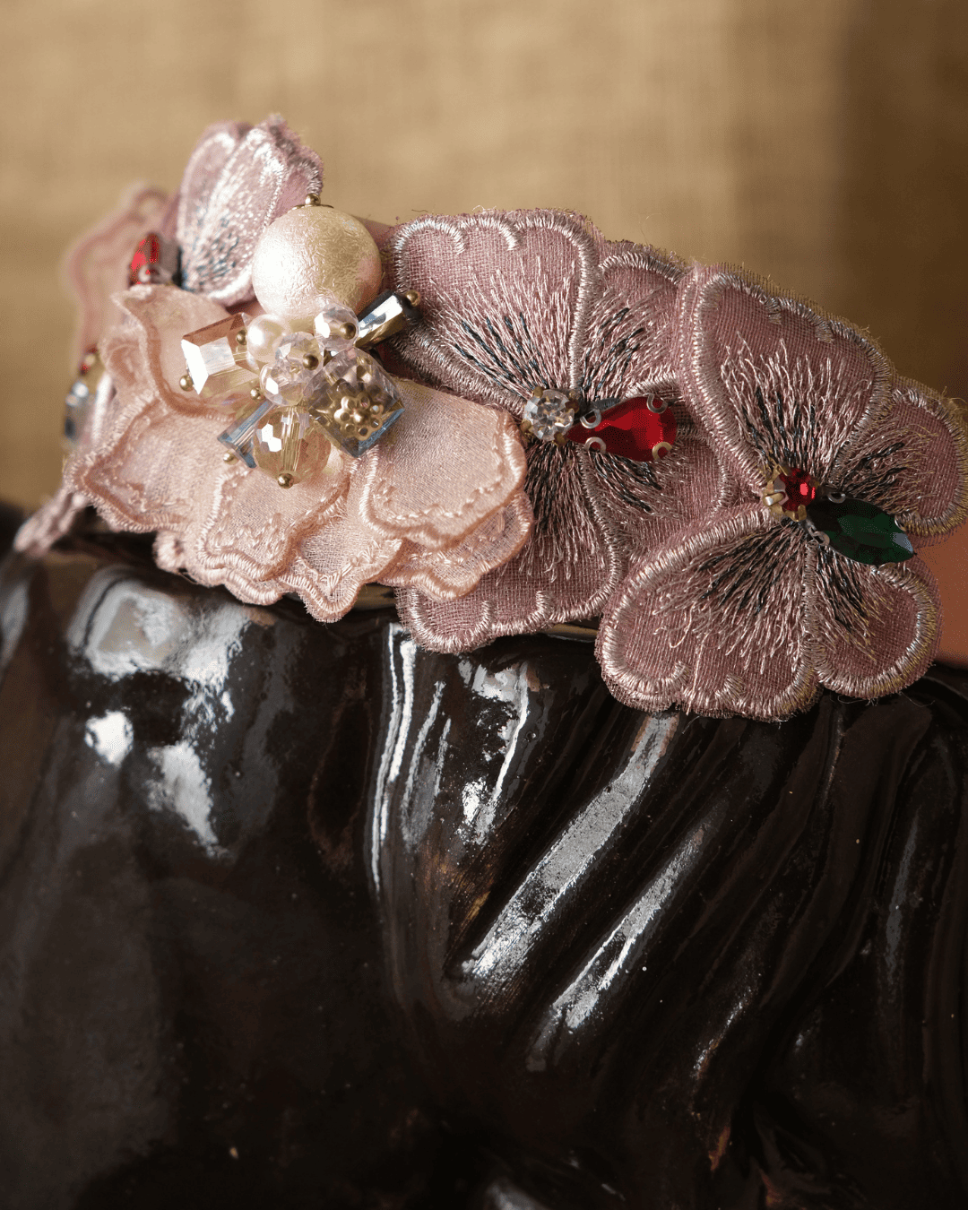 A close-up of a decorative headband featuring layered dusty pink fabric flowers, rhinestone embellishments, and a pearl-like bead, displayed on a glossy dark surface against a warm beige background.