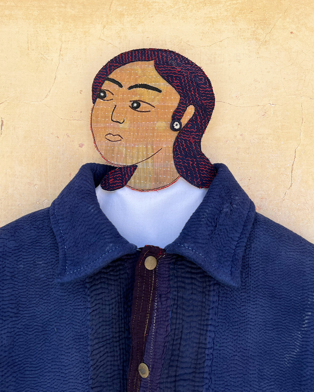 Textile art piece of a person wearing a blue coat with a textured head and hair. day-and-age-marina-jacket-two-extra-lives