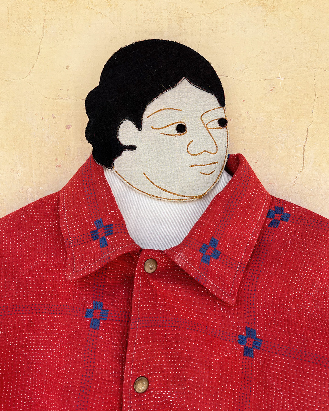 Textile art piece of a person wearing a red shirt with blue patterns on a beige background day-and-age-steaam-angine-jacket-two-extra-lives