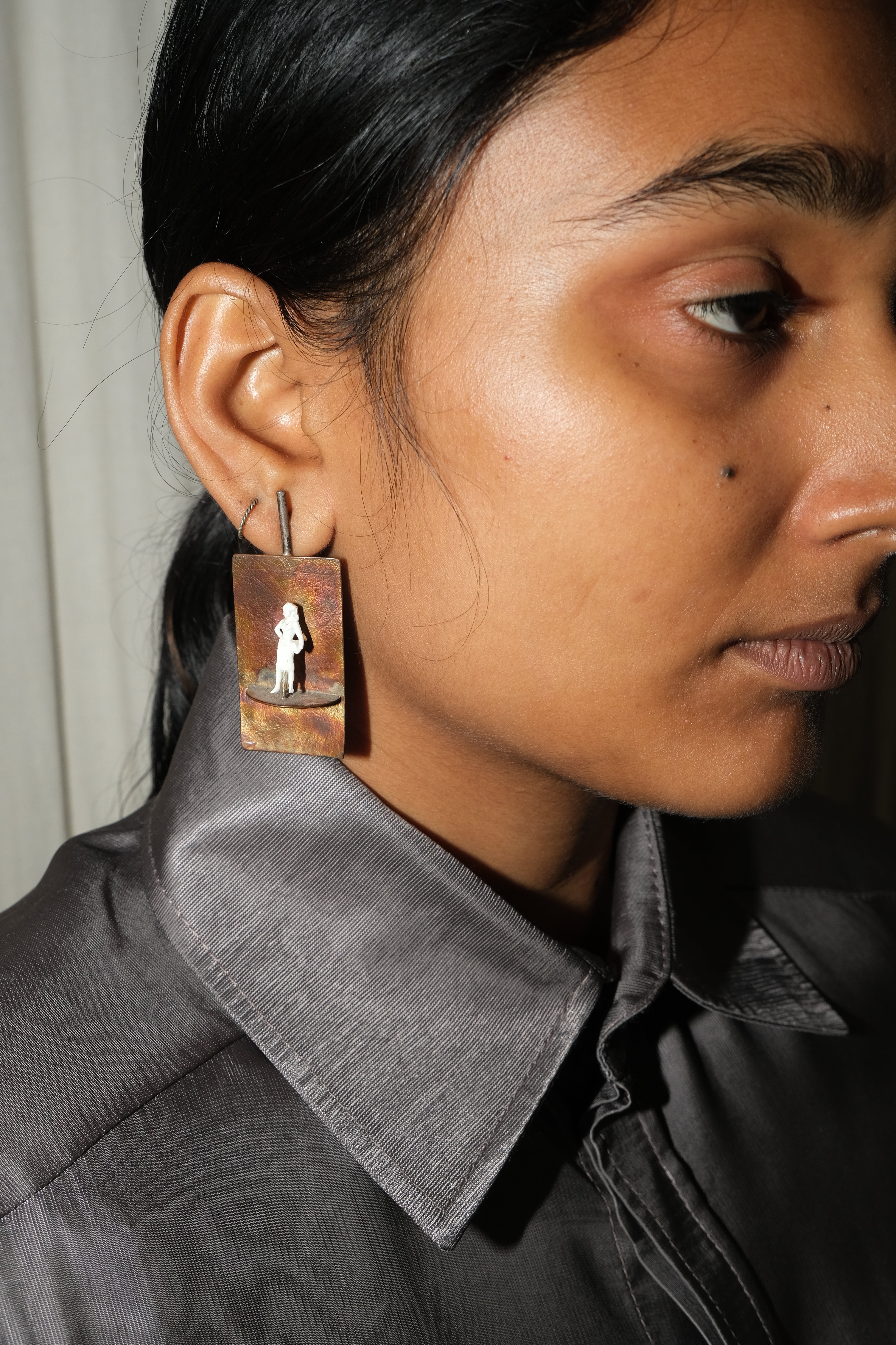 A close-up side profile of a person wearing a rectangular brown brass earring with a small white silhouette figure printed on it, paired with a dark gray collared shirt.
