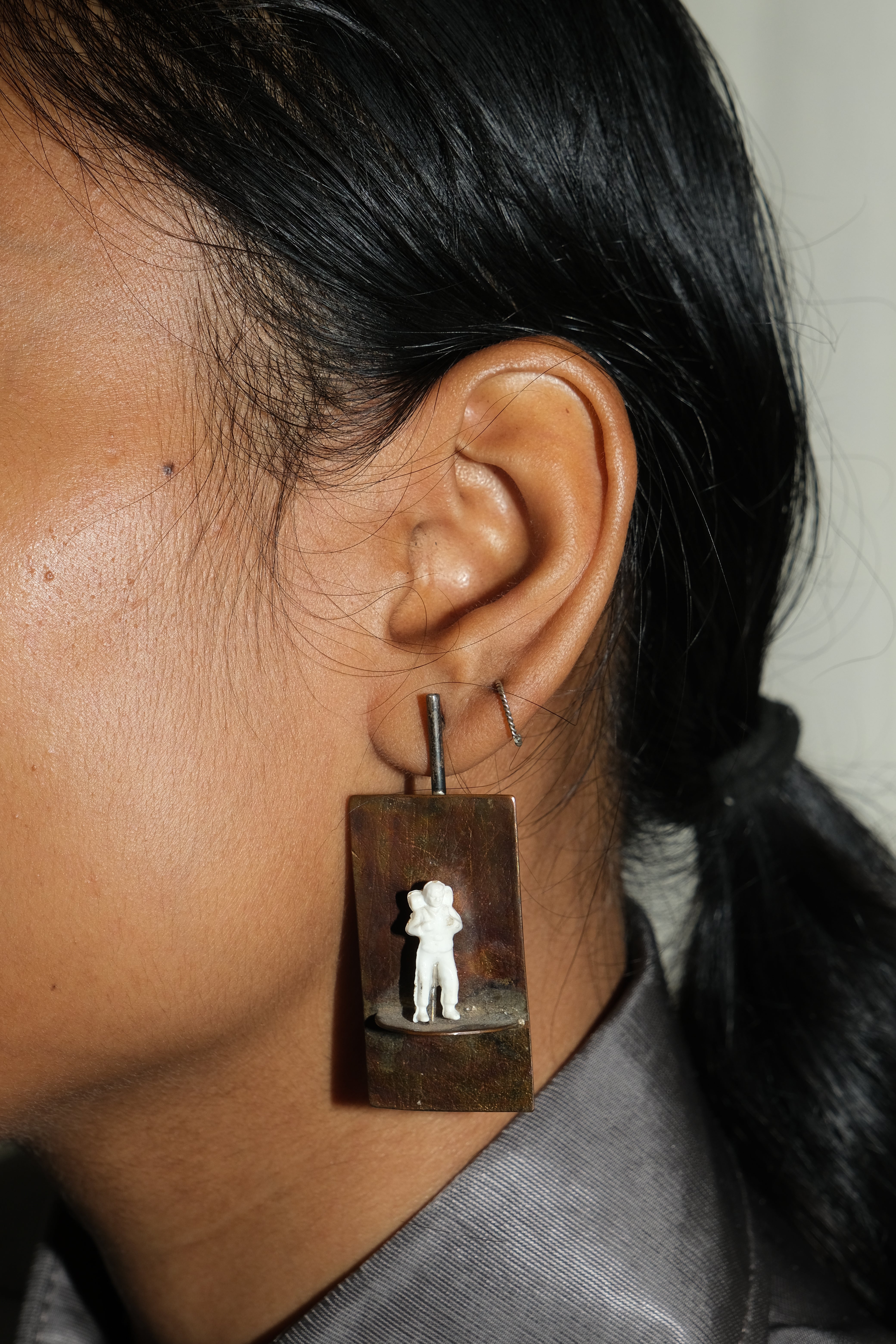 A close-up side profile of a person wearing a large rectangular drop earring. The earring has a dark brown wood or metal frame with a small white figurine of two people standing together, and a thin silver hoop above it.